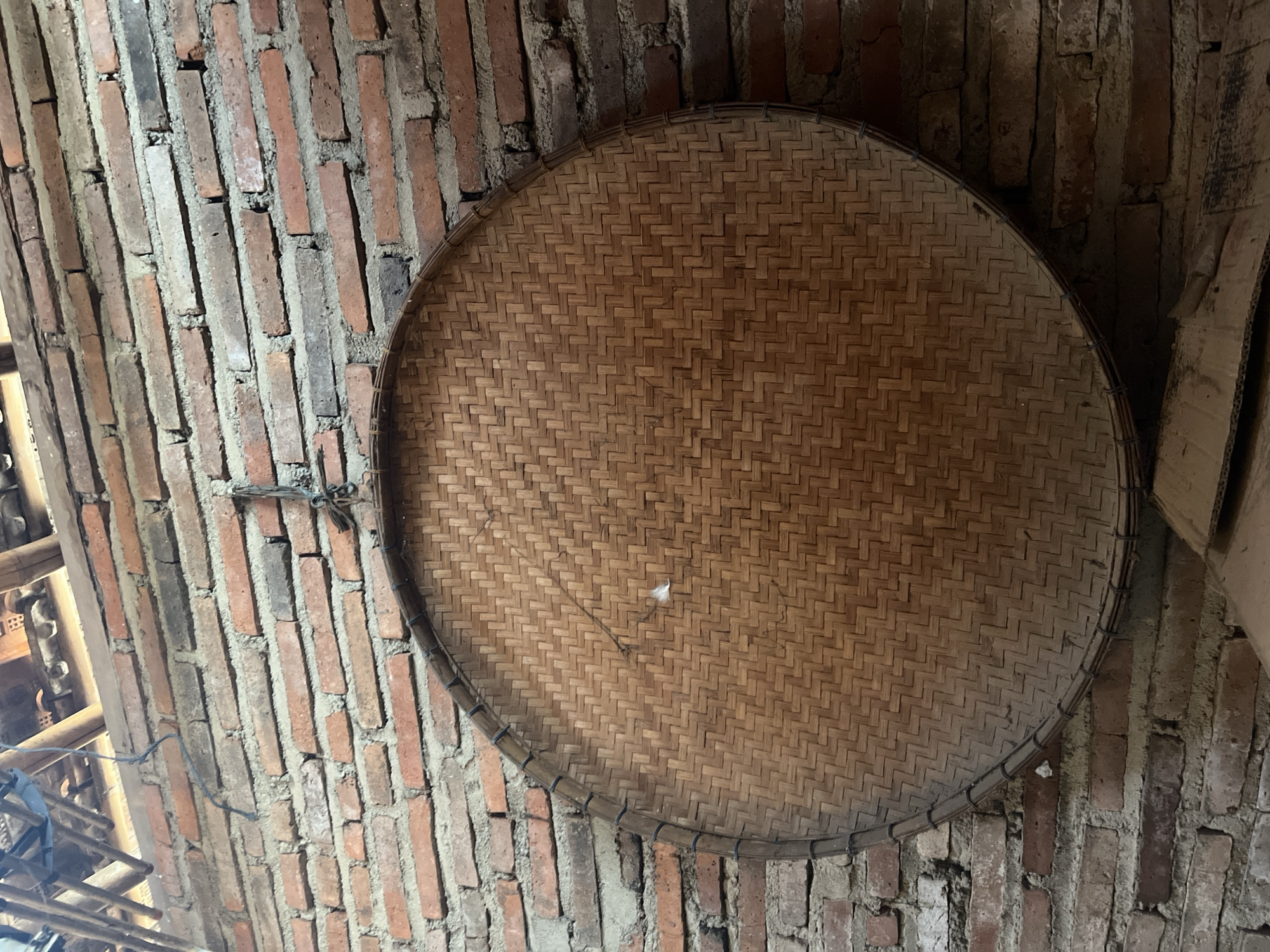 A large, round, woven bamboo tray is hanging on a red brick wall. The tray has a woven pattern of diagonal lines, forming a lattice. The brick wall is made of rough, uneven bricks, some of which are a reddish-brown color, and others are darker, appearing almost black. The mortar between the bricks is visible. Above the tray and wall, parts of a wooden ceiling structure and roofing material are visible, suggesting an indoor or semi-outdoor setting. The overall impression is of a rustic, possibly agricultural, environment. The location is specified as Glagah Kulon, Indonesia.