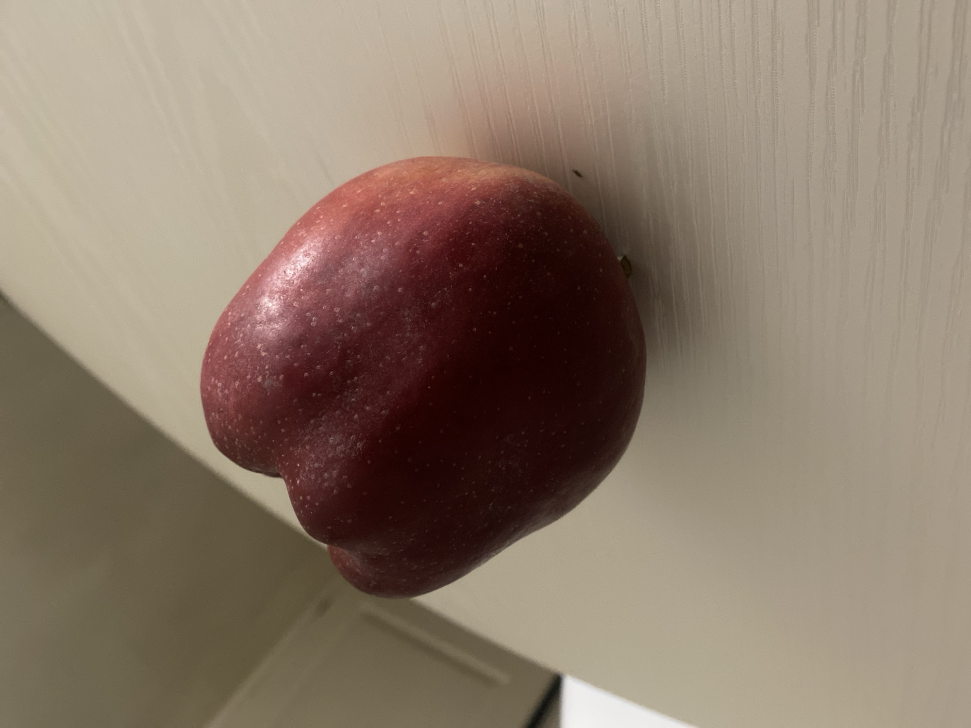 A red apple with an unusual, lumpy shape rests on a textured, light-colored surface. The surface appears to be a table or countertop with vertical indentations. The lighting in the scene is dim, casting a shadow from the apple onto the surface. The background is out of focus and appears to be a neutral-colored wall or cabinet. There are no people or other objects visible in the image.