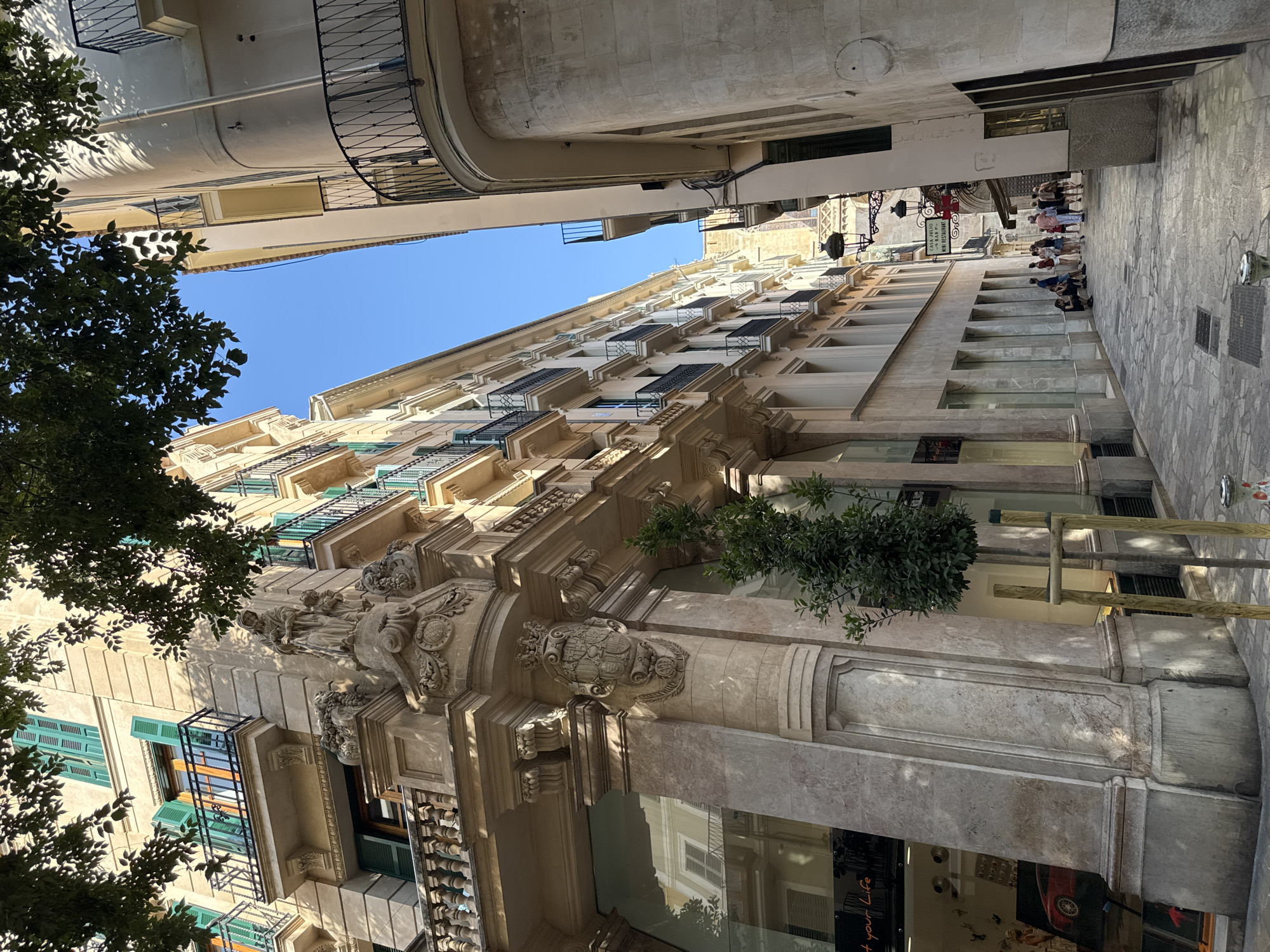The image depicts a narrow street in Palma, Spain, with historic buildings flanking both sides. The architecture features ornate facades with balconies, arched windows, and decorative stonework, suggesting an older European urban environment. Lush green trees with visible foliage cast shadows on the buildings and the cobblestone street. The sky is a clear, bright blue.
On the left, a building exhibits a classical style with carved stone details above a storefront. A modern element is present with a large glass window revealing displays inside, including what appears to be a red car. A young tree is planted on the sidewalk next to the building, supported by wooden stakes.
On the right, another building presents a curved facade, also with balconies. Further down the street, a group of people, appearing to be tourists, are gathered and walking. They are casually dressed, consistent with a warm climate. A street lamp with intricate metalwork is visible, along with a sign for "CASA JULIO BAR & RESTAURANT". The overall atmosphere is one of a sunny day in a vibrant, historical city.