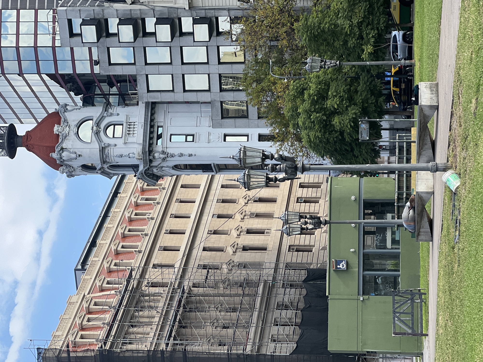 This vibrant daytime image captures a diverse urban streetscape in Buenos Aires, Argentina, under a clear sky with scattered white clouds. The scene is dominated by a blend of architectural styles and daily city life.
In the midground, an ornate, light-colored building stands out with a distinctive red dome or cupola and decorative classical elements, including arched and round windows. To its right, a modern, tall building features a grey facade with numerous reflective, square windows, some of which protrude, mirroring the sky. To the left, an older, multi-story beige building is partially obscured by extensive scaffolding and black netting, indicating renovation or construction work. This building has repetitive window designs and decorative cornices with reddish accents.
In the foreground, a strip of manicured green grass and a paved concrete walkway separate the viewer from the road and buildings. Several traditional multi-lamp streetlights are positioned along the walkway. A low, rectangular green building or structure is visible on the right, featuring large windows with faint graffiti-like text and a prominent wheelchair accessibility symbol.
On the street behind the grass, a yellow and black taxi is parked, and a silver car is also visible. Part of a green and yellow bus, bearing the number "7033," can be seen further down the road. A person is lying or sitting on a concrete bench-like structure near the green building, and another person is partially visible near the taxi. The mix of classic and modern architecture, along with ongoing construction and public transport, depicts a dynamic and evolving urban environment.