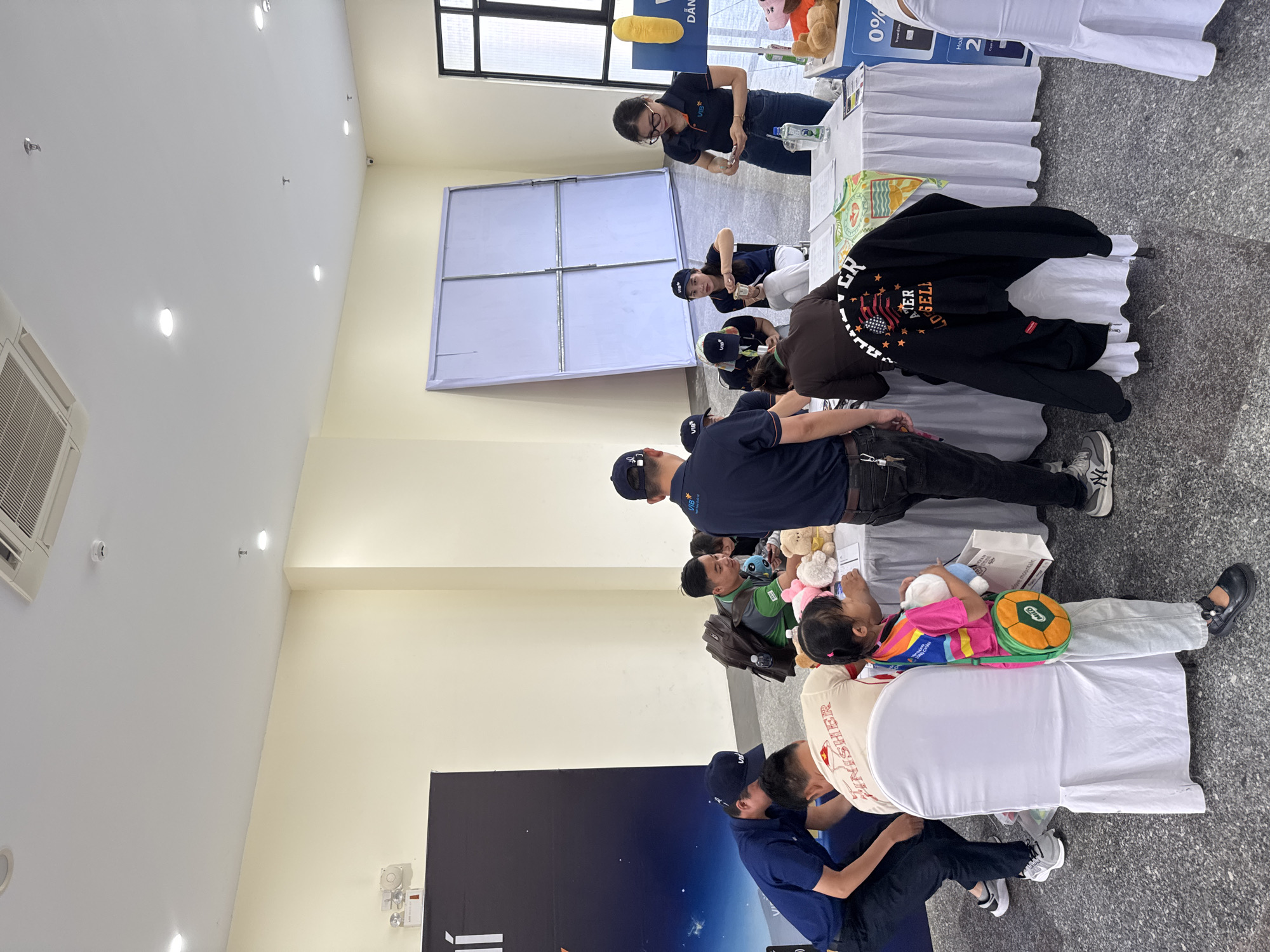 The image depicts an indoor event, likely a promotional booth or registration area, set against a neutral, off-white wall. A large black banner with white lettering is visible on the left. In the center, a white-covered table is laden with papers, a water bottle, and plush toys. Several individuals are present. On the left, a person in a dark blue shirt and cap is bent over the table. To their right, a person in a cream-colored t-shirt with red lettering is facing away from the camera. A young girl in a brightly colored shirt and a green and orange circular purse stands near the table, holding a plush toy. Further to the right, a group of people are gathered around the table. One person in a dark blue shirt and cap is holding a smartphone, and another individual is handling money. A woman in a dark blue shirt and glasses is also at the table, appearing to count money. In the background, a large white board leans against the wall. The floor is a speckled gray material. No specific Hoi An landmarks or indications of the city are visible.