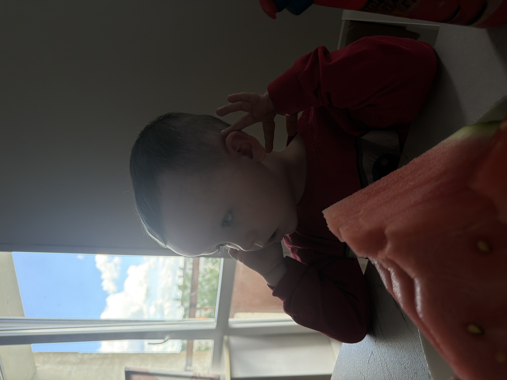 A young child is seated at a light-colored table indoors in the city of Chaikovsky, Russia. The child, wearing a red long-sleeved shirt, is depicted in profile, facing left with a slightly open mouth. Their left hand is raised, with fingers positioned near their left ear, and their right hand is similarly raised near the back of their head. A large slice of watermelon, displaying reddish-pink flesh, several dark seeds, and a green rind, occupies the foreground on the table. A prominent window on the left side of the frame reveals an outdoor daytime scene consisting of a bright blue sky with white cumulus clouds, green foliage, and what appears to be a distant building. The interior lighting is uneven, with strong natural light from the window contrasting with a darker background behind the child. A red bottle is partially visible on the far right side of the table.