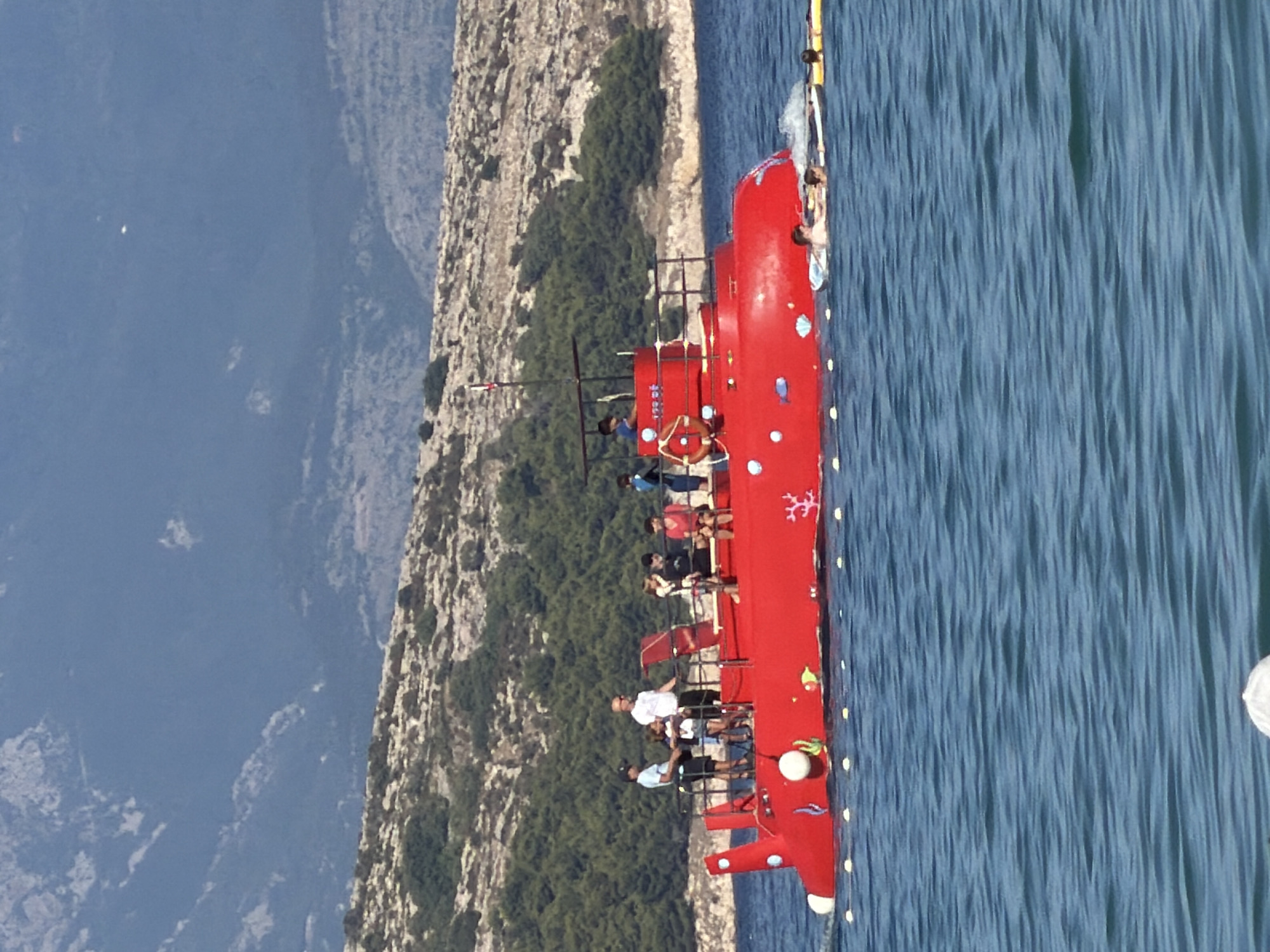 A red submarine-shaped tourist boat is on the water in Općina Baška, Croatia, with people standing on its deck and swimming nearby. The boat is adorned with cartoonish sea creatures and a life preserver. The background features a rocky, vegetated hillside and a hazy mountain range under a clear sky. The water is calm and blue.