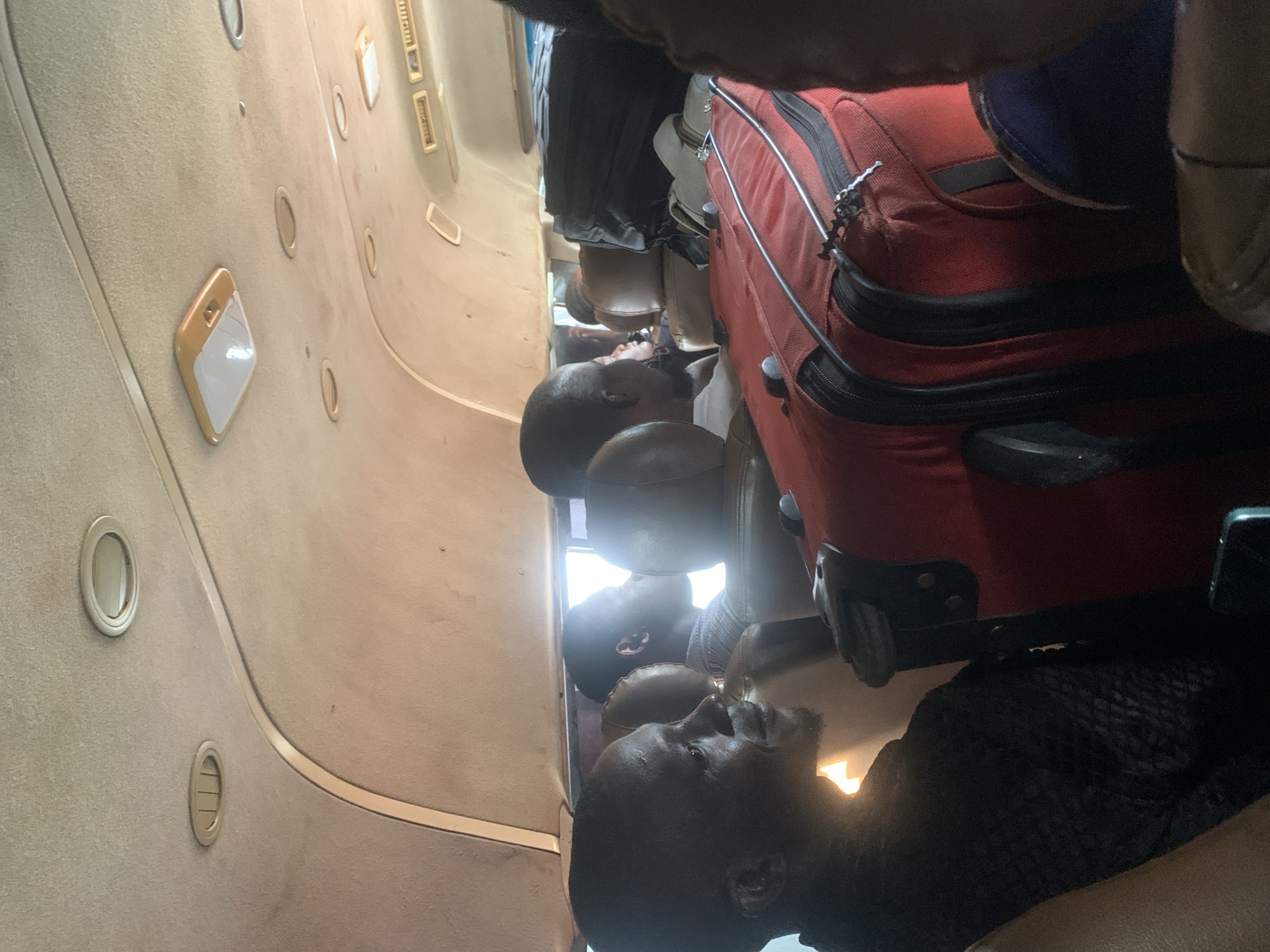 The image shows the interior of a passenger van, likely a commercial bus, with several individuals seated inside and luggage packed around them. The ceiling of the van features overhead lighting and air vents. The passengers appear to be of African descent. One passenger in the foreground is seen in profile, looking forward. A large red suitcase is prominently placed in the aisle, alongside other dark-colored luggage. The scene suggests travel, possibly a long-distance journey. The image context provided states the location is Elelenwo, Nigeria.