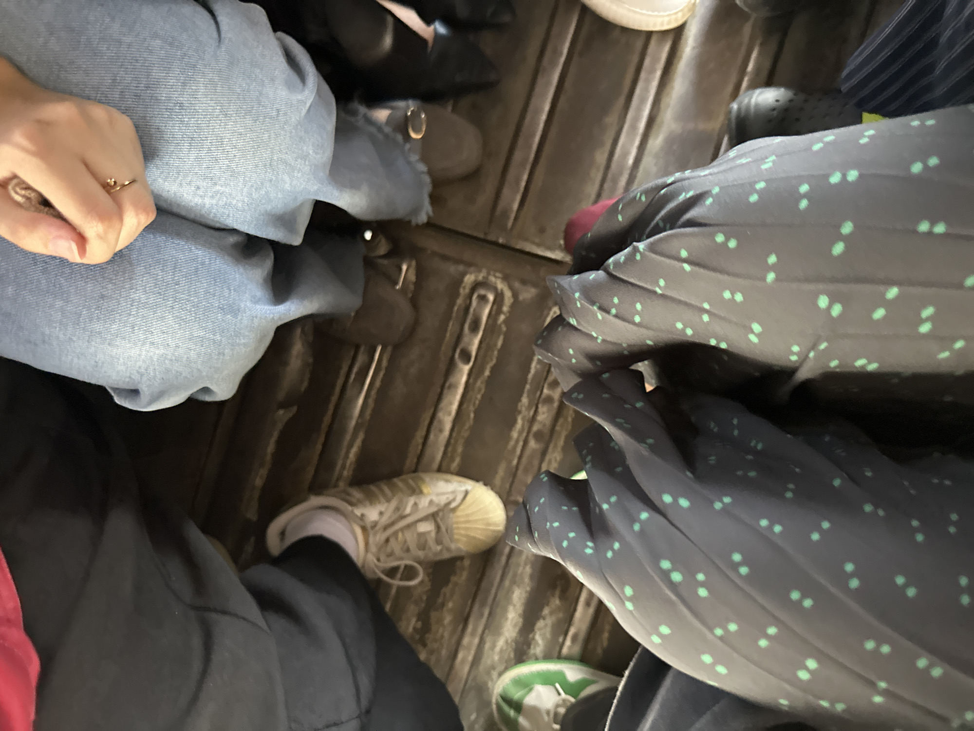 The image shows a view of people's legs and shoes on what appears to be the floor of a bus or similar public transport in Davao City, Philippines. The floor is ridged, with multiple pairs of shoes and feet visible. One person wears light-wash denim jeans. Another person wears dark trousers and off-white sneakers with white socks. A third person is wearing a dark-colored dress or pants with small light green polka dots, and another person wears white sneakers with green markings. At least one person has a ring on their finger. The image lacks any information identifying the specific location.