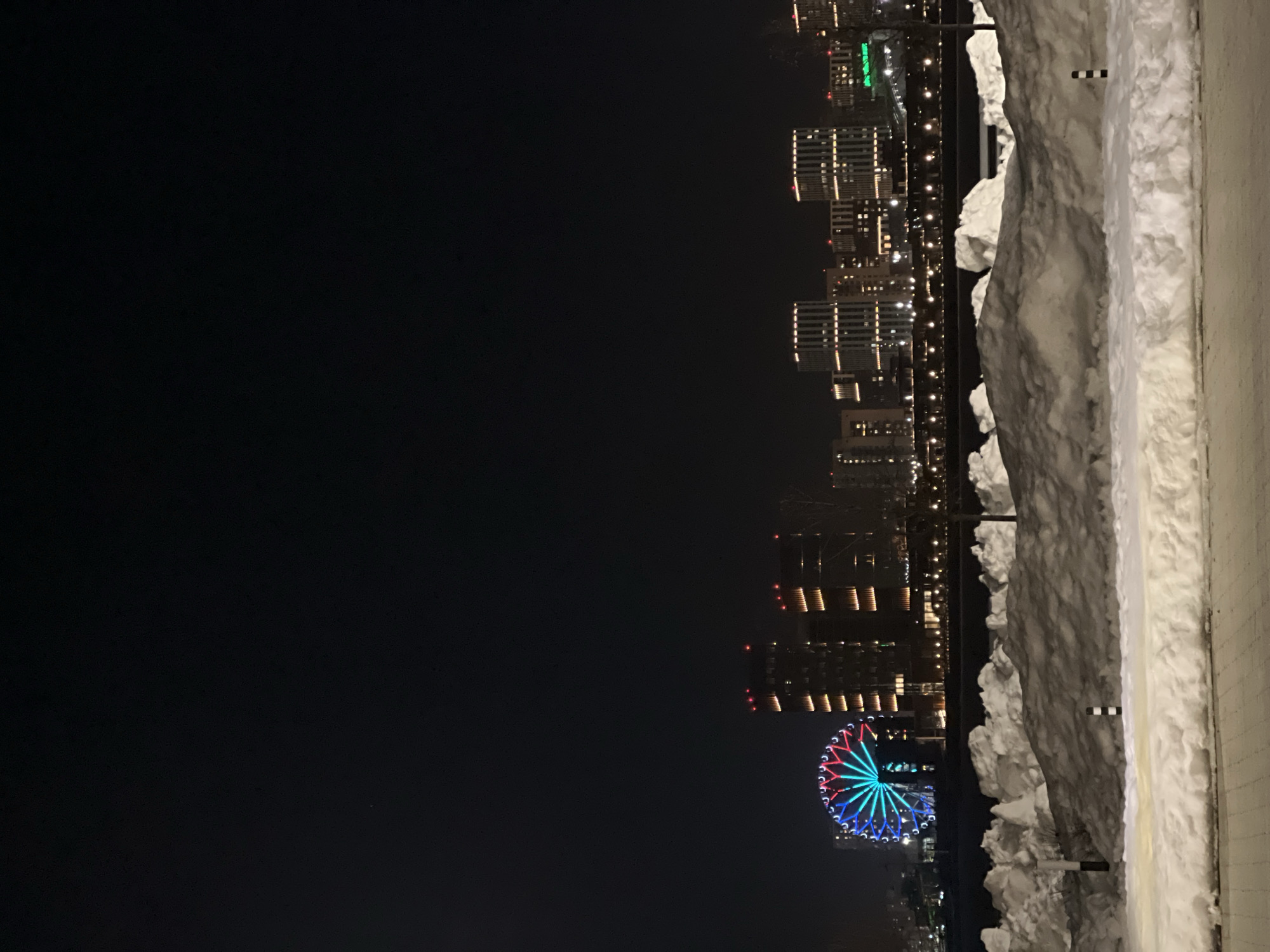 The image displays a nighttime winter cityscape in Kazan, Russia, likely viewed across the Kazanka River towards the Riviera complex. In the immediate foreground, a paved surface transitions into an extensive, uneven bank of snowdrifts, exhibiting shades of white and gray. Two small, black and white striped bollards are partially visible within these snowdrifts. Beyond the snow, a darker expanse, possibly a frozen waterway, separates the foreground from the distant city lights. The midground features a linear arrangement of small, bright lights, suggesting a bridge or waterfront promenade.
The background reveals a densely illuminated urban skyline. On the left, a large Ferris wheel, known as the Kazan Ferris Wheel (Kazan Eye), stands prominently, illuminated with segments of red, white (or light blue), and blue lights. To its right and extending across the horizon are numerous multi-story buildings, characterized by regular patterns of warm white or yellow window and facade lighting, with some rooftops displaying intermittent red lights. No individuals are visible, and no dynamic actions or interactions are depicted within the scene, which is static.