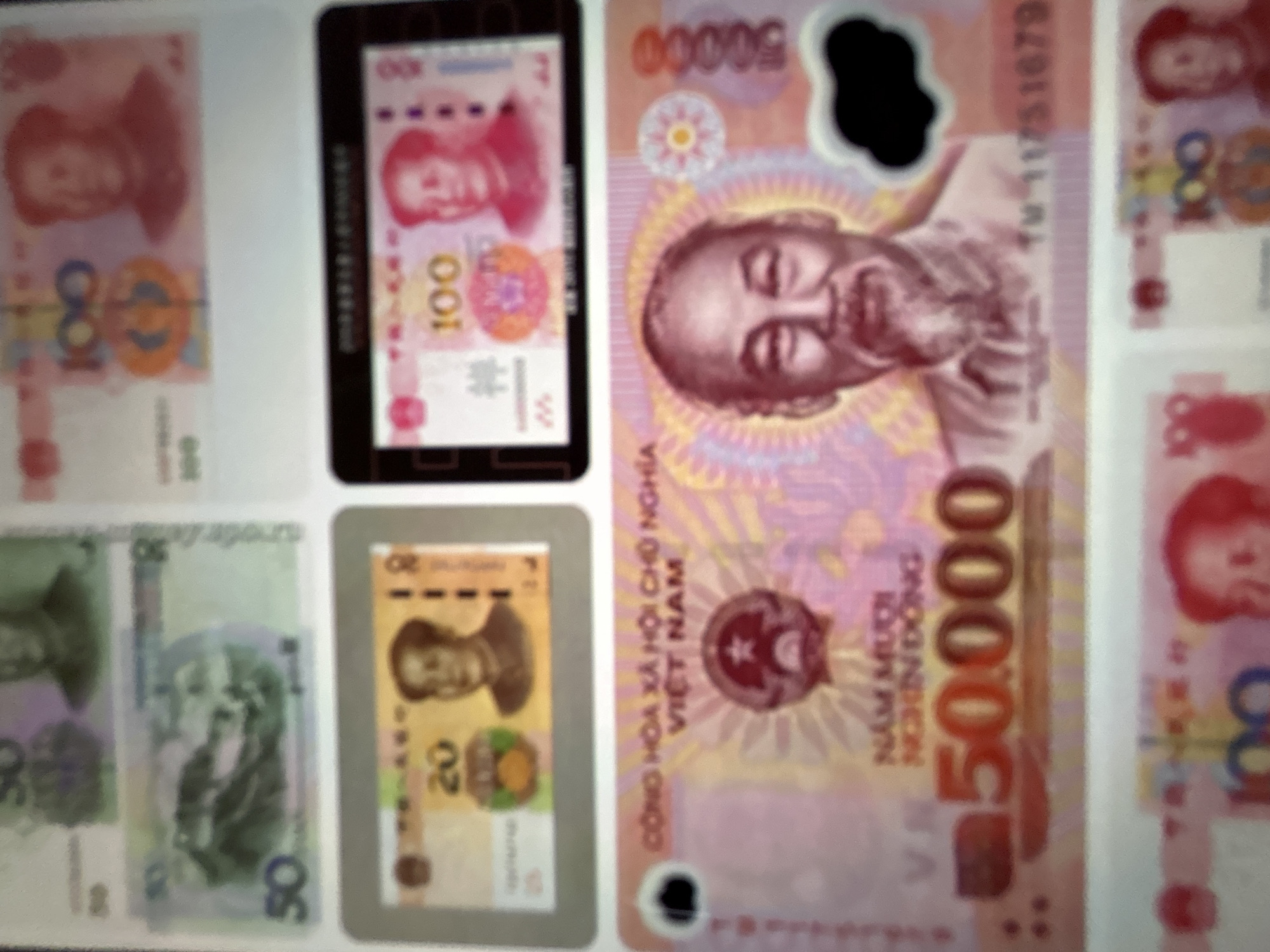 This media file showcases a collection of banknotes, primarily focusing on currencies from China and Vietnam. The banknotes are arranged in a display, possibly for educational or collection purposes.
On the left, several Chinese Yuan banknotes are visible. One clearly displays the denomination "100" in large numerals and features a portrait of Mao Zedong. Another partially visible banknote also appears to be a "100" Yuan note. Below these, a lighter colored banknote with the denomination "50" is present.
To the right of the Chinese currency, a Vietnamese Dong banknote stands out. It is predominantly pink and purple, with a large "50,000" denomination prominently displayed. The portrait of Ho Chi Minh is clearly visible on this banknote. Text in Vietnamese, including "CÔNG HÒA XÃ HỘI CHỦ NGHĨA VIỆT NAM," is also legible.
Further to the right, more Chinese Yuan banknotes are visible, including another "100" denomination note.
The background is plain white, suggesting the banknotes are laid out on a flat surface. The lighting appears to be artificial and relatively bright. There are no discernible details about the time of day, weather, or specific location beyond the currencies themselves. The focus is entirely on the financial instruments, with no discernible activity or event taking place other than the presentation of the banknotes. The image is slightly blurry, making precise identification of all details challenging.
