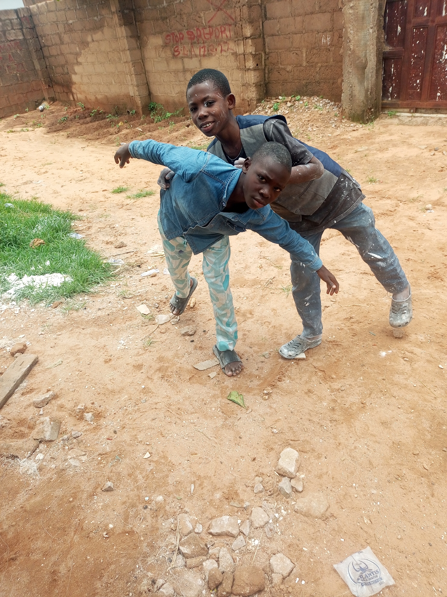 Two young boys, appearing to be of African descent, are posing for a photograph in what seems to be an outdoor, urban environment. The boy in the foreground, wearing a denim jacket and patterned pants, is leaning forward and looking directly at the camera with a playful expression. The boy behind him, in a dark vest over a t-shirt and dark pants, has his arm around the other boy's shoulders, also looking towards the camera. Both boys have white paint or dust splattered on their faces and clothing, suggesting they might have been involved in some sort of construction or artistic activity.
The background consists of a brick wall with graffiti, and a weathered wooden door. The ground is sandy and uneven, with scattered rocks and some patches of grass. A plastic water sachet is visible on the ground in the lower right corner. The overall impression is of a candid snapshot taken in a somewhat unkempt, but lively, urban setting in Tempure, Nigeria.