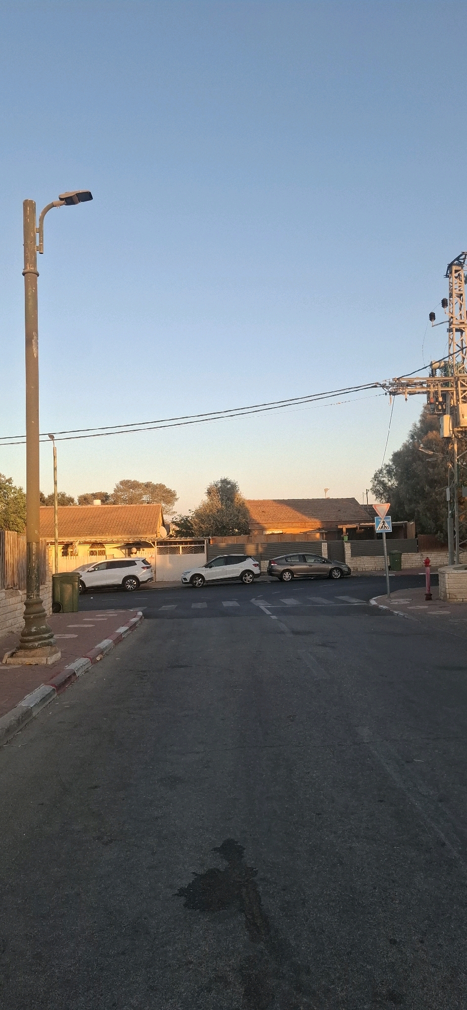 In Yerocham, Israel, a street scene unfolds under a clear, pale blue sky during what appears to be early morning or late afternoon, indicated by the warm, golden light illuminating the rooftops. A prominent, weathered lamppost stands on the left, its dark metal contrasting with the bright sky. Adjacent to it, a green trash bin sits on a sidewalk bordered by red and white striped curbing. The street is paved with asphalt, showing some wear and a dark, irregular stain in the foreground.
Parked along the right side of the street are three vehicles: a white SUV on the far left, followed by another white SUV, and a darker, possibly gray or brown SUV on the far right. Behind the cars, residential buildings with reddish-brown tiled roofs are visible, interspersed with trees and foliage. A dark fence separates the properties from the street.
Further down the street, on the right, a utility pole with electrical wires and equipment stands tall. A diamond-shaped pedestrian crossing sign with a blue background and a white silhouette of people walking is also visible near the intersection. A red fire hydrant is positioned on the corner. The overall impression is a quiet, residential street in a small Israeli city, bathed in soft, directional light.