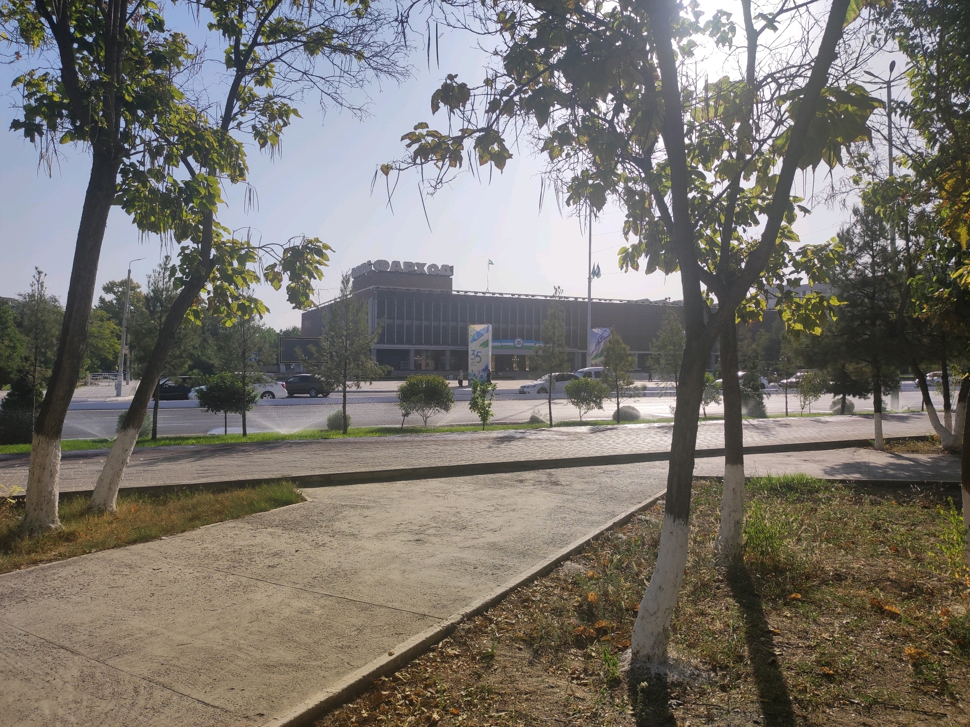 The image depicts a view of a building in Navoiy City, Uzbekistan. The building is a low-rise structure with a facade featuring multiple windows and prominent lettering on its roof that appears to be in Cyrillic script. In front of the building, there is a street with cars, and trees line the sidewalk. The foreground shows a paved walkway and grassy areas with scattered leaves, indicating an outdoor environment. The sky is clear and bright, suggesting daytime. The overall scene appears to be a public space or a commercial area within the city.