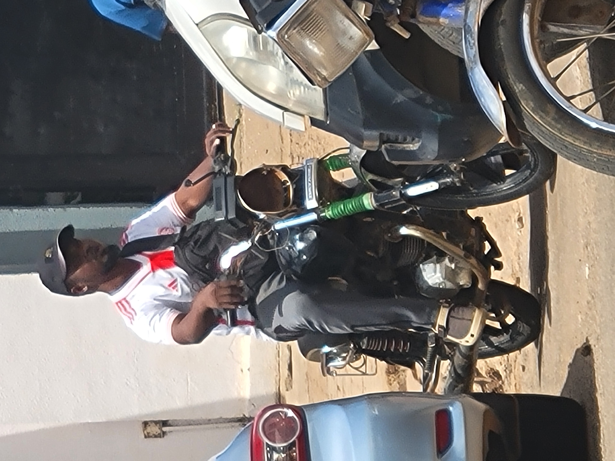 A man wearing a white t-shirt with red stripes, gray pants, a black cap, and a dark bag slung over his shoulder sits on a motorcycle in an outdoor setting, potentially in Jos, Nigeria. He appears to be manipulating the motorcycle's controls. The motorcycle is between a white vehicle and a blue vehicle. The photo is taken at a slight angle, with the buildings and vehicles appearing tilted.