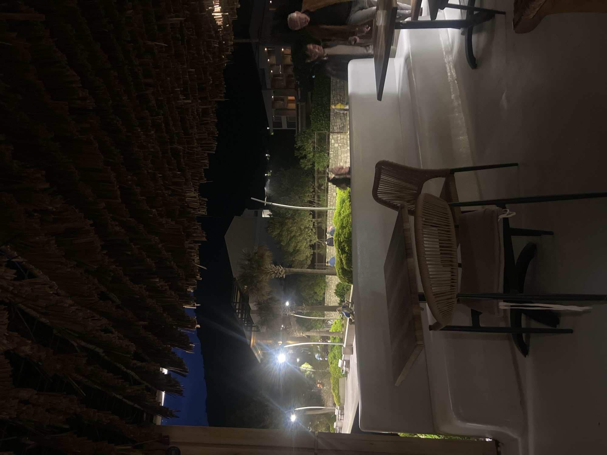 Nighttime scene at a venue in Petrovac centar, Montenegro. A thatched roof covers the foreground, while chairs and tables occupy the immediate area. People are seated at one of the tables. Beyond the seating area, the scene opens to reveal a walkway illuminated by lights, lined with trees and vegetation. Buildings are visible further in the background. The sky is dark.