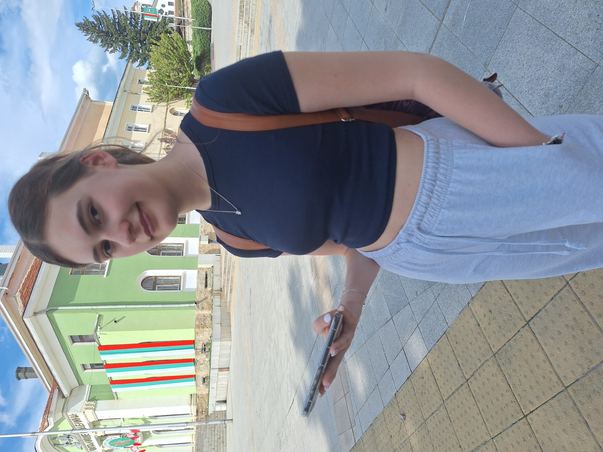 A young woman takes a selfie outdoors, holding a smartphone in her right hand. She is wearing a dark blue crop top, light gray sweatpants, and a brown shoulder bag. Her face is turned towards the camera with a slight smile. In the background is a green and white building with horizontal stripes of white, red, and green, resembling the Bulgarian flag. The building is identified as a government office in the city of Panagyurishte, Bulgaria, indicated by the Cyrillic script and flag colors. The background also shows portions of other buildings and a tree under a partly cloudy sky. The scene is on a paved walkway.