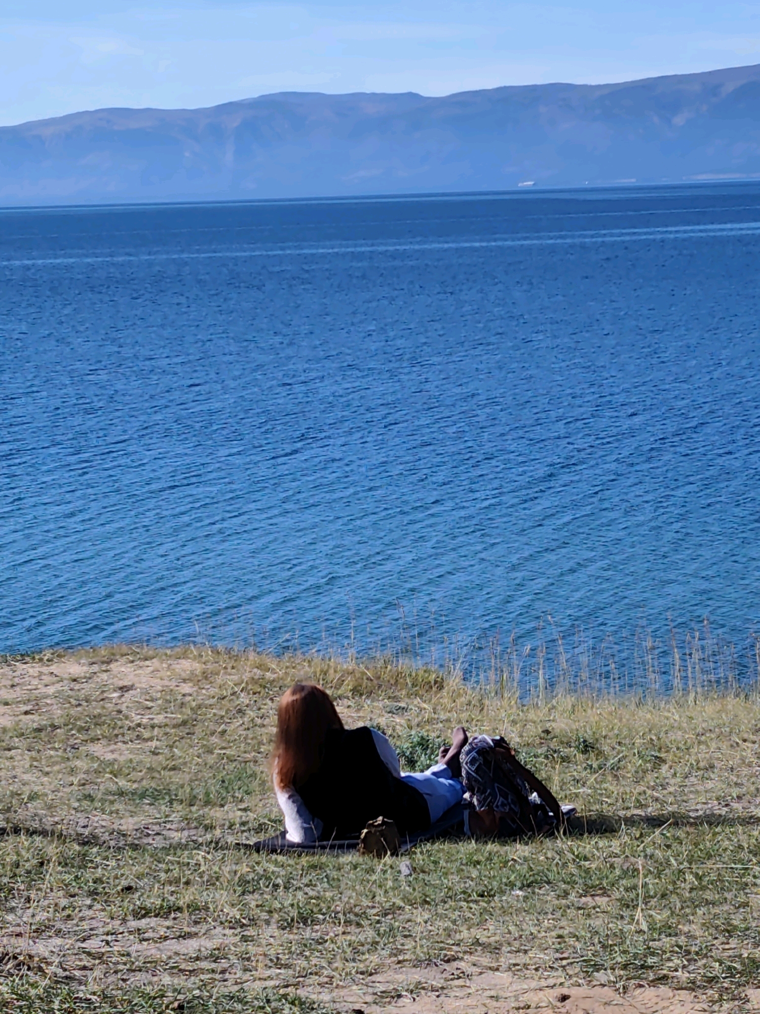 A person with long, reddish-brown hair lies on their back in the grass on a grassy embankment, facing away from the camera. They are wearing a light-colored shirt and dark pants, with their legs crossed. A backpack is placed next to them on the grass. The background features a wide expanse of blue water with gentle ripples, suggesting a lake or sea. In the distance, a range of hazy, blue mountains forms the horizon, under a light blue sky with some clouds. The scene appears tranquil and offers a sense of natural beauty and relaxation. This is a view of Lake Baikal, near the village of Kharantsy in Russia.