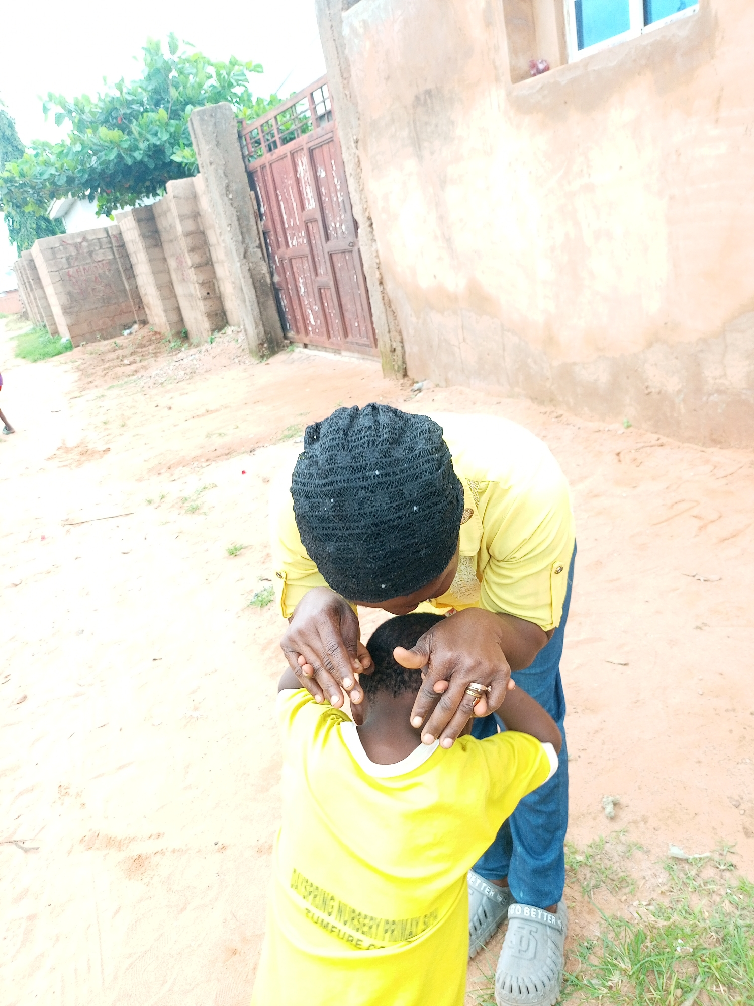 The image shows two people, an adult and a child, outdoors in what appears to be Tempure, Nigeria. The adult, wearing a yellow shirt and a black headscarf, is bending down to gently hold the back of the child's head. The child, facing away from the camera, is wearing a bright yellow t-shirt with white trim. The t-shirt has visible text that reads "DISPRING NURSERY PRIMARY SCH TUMFURE".
The setting is a sandy, dirt path. In the background, there is a rustic wall made of concrete blocks and bricks, with a weathered metal gate. To the left of the gate, there are green trees and foliage. To the right, a building with a light-colored, textured wall is visible, featuring a window with blue panes. The ground is a mix of sand, dirt, and some sparse green grass. The lighting suggests it is daytime, possibly late morning or early afternoon. The scene appears to be casual and intimate, with the adult's gesture suggesting care or affection towards the child. The overall impression is of a simple, everyday moment in a Nigerian setting.