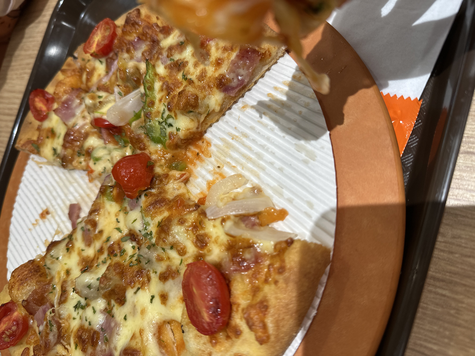 The image shows a partially eaten pizza on a brown-rimmed plate. The pizza has a topping of cheese, tomatoes, onions, and what appears to be ham or bacon. The plate rests on a black tray. The location is Chongqing High-tech Zone, China, suggested by the context of the provided query. The angle and lighting suggest the photo was taken indoors and recently. There is a pizza slice being lifted.