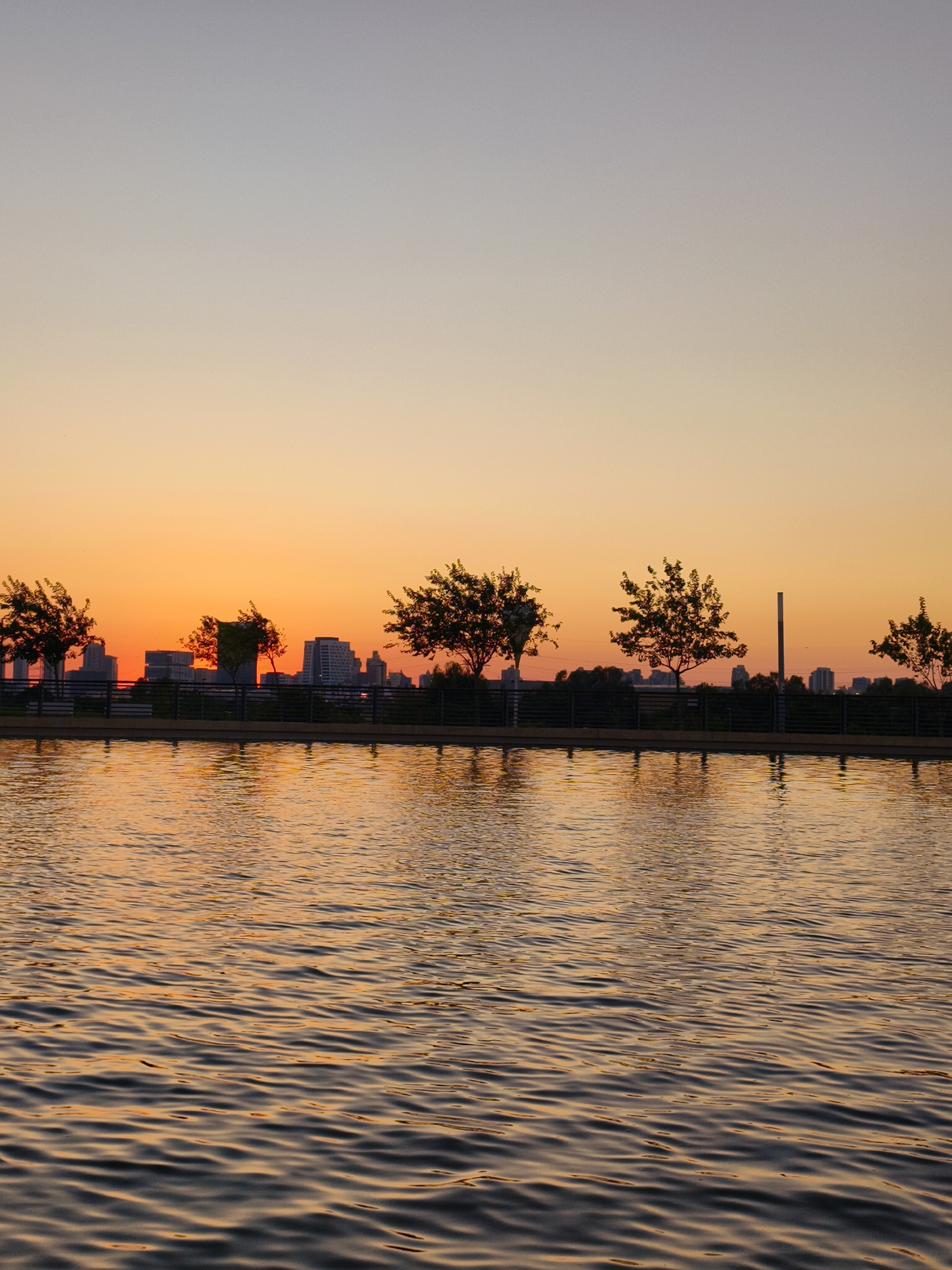 This is a photograph of the sunset over the Beer Sheva River Park in Be'er Sheva, Israel. The sky transitions from a pale blue at the top to a vibrant orange near the horizon. Silhouetted trees line the bank of a body of water in the foreground, which reflects the colors of the sunset. In the background, a cityscape with buildings is visible, also silhouetted against the sky. The water is rippled, creating a textured surface.