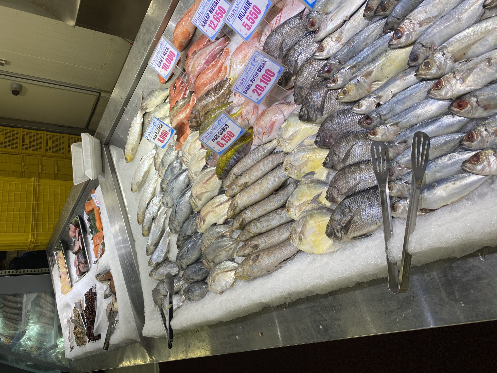 This image shows a fresh fish market stall in Banda Aceh, Indonesia. The stall is filled with various types of fish displayed on ice. Several price tags are visible, indicating different varieties of fish and their prices in what appears to be Indonesian Rupiah. In the background, yellow crates are stacked, commonly used for storage and transport in markets. The overall scene depicts a bustling marketplace offering a fresh selection of seafood.