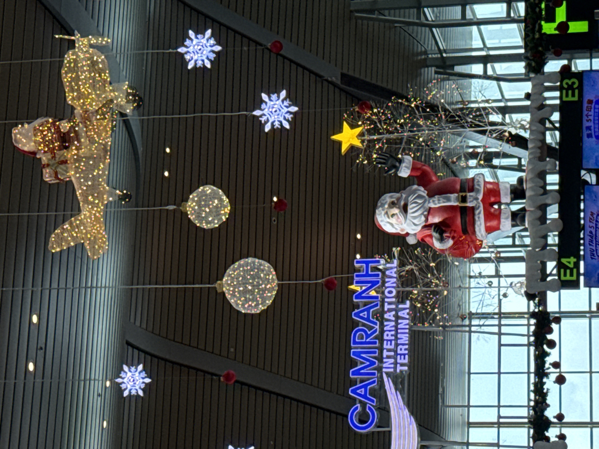 The image shows a festive Christmas display at Cam Ranh International Terminal in Cam Ranh, Vietnam. A large, illuminated Santa Claus figure is positioned prominently, waving with his right hand. Behind Santa, a decorated Christmas tree with colorful lights is visible. Overhead, an illuminated airplane sculpture with Santa Claus riding on top is suspended, along with several glowing spherical ornaments and snowflake decorations. The terminal's signage, reading "CAMRANH INTERNATIONAL TERMINAL," is clearly visible in blue illuminated letters. The background consists of large windows looking out onto an outdoor area, and the interior ceiling of the terminal with its slatted design. Various Christmas-themed decorations, including garlands and red ornaments, are also present. The overall scene captures a holiday ambiance within an airport setting.