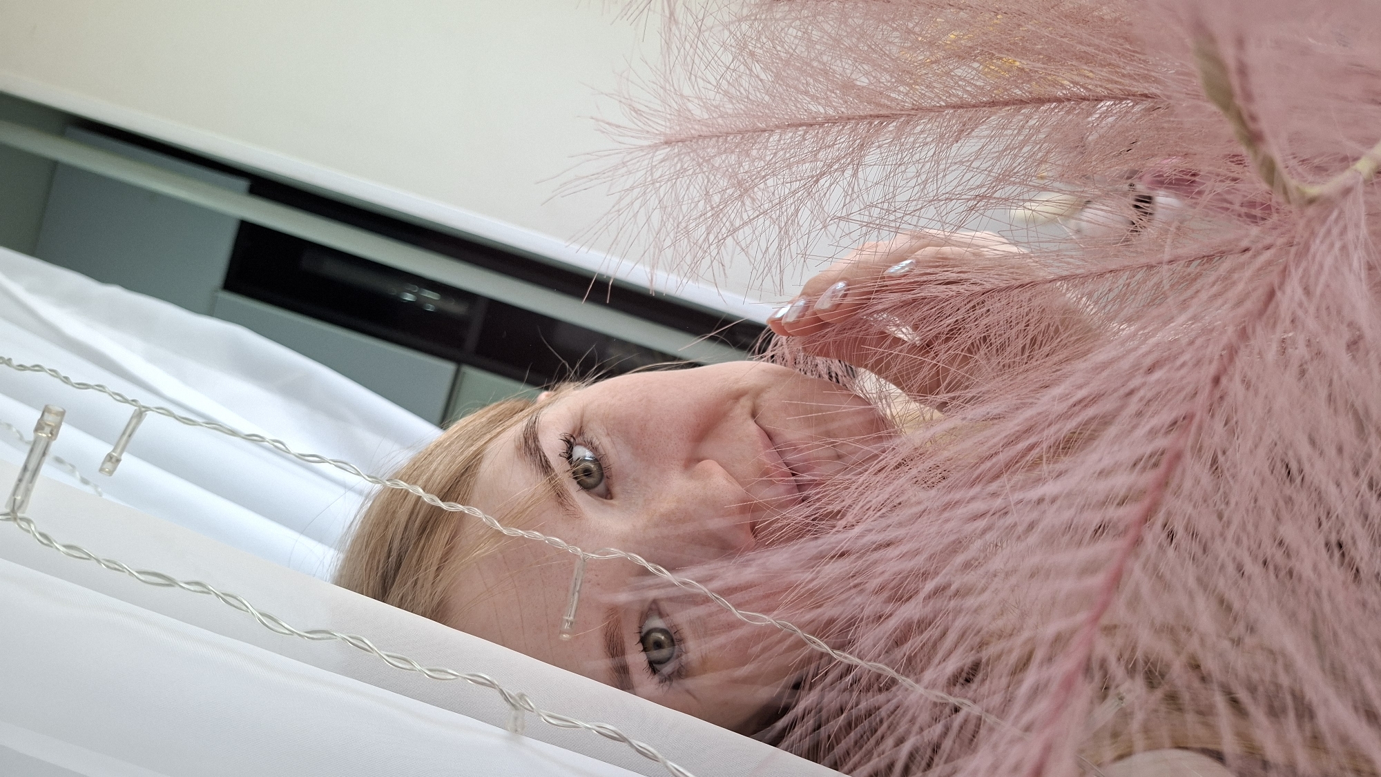 The image shows a young woman with blonde hair and fair skin, likely lying down, as indicated by the tilted perspective and the items above her. Her face is visible between strings of small LED lights and a large bundle of fluffy, pink Pampas grass. She has light green eyes and freckles on her face. Her hand, with long, light-colored nails, is partially visible near the Pampas grass. Above her is a white surface and what appears to be a built-in unit with a dark horizontal element. The location is presumed to be in Chaikovsky, Russia, though there are no specific landmarks or objects in the image to confirm the location.