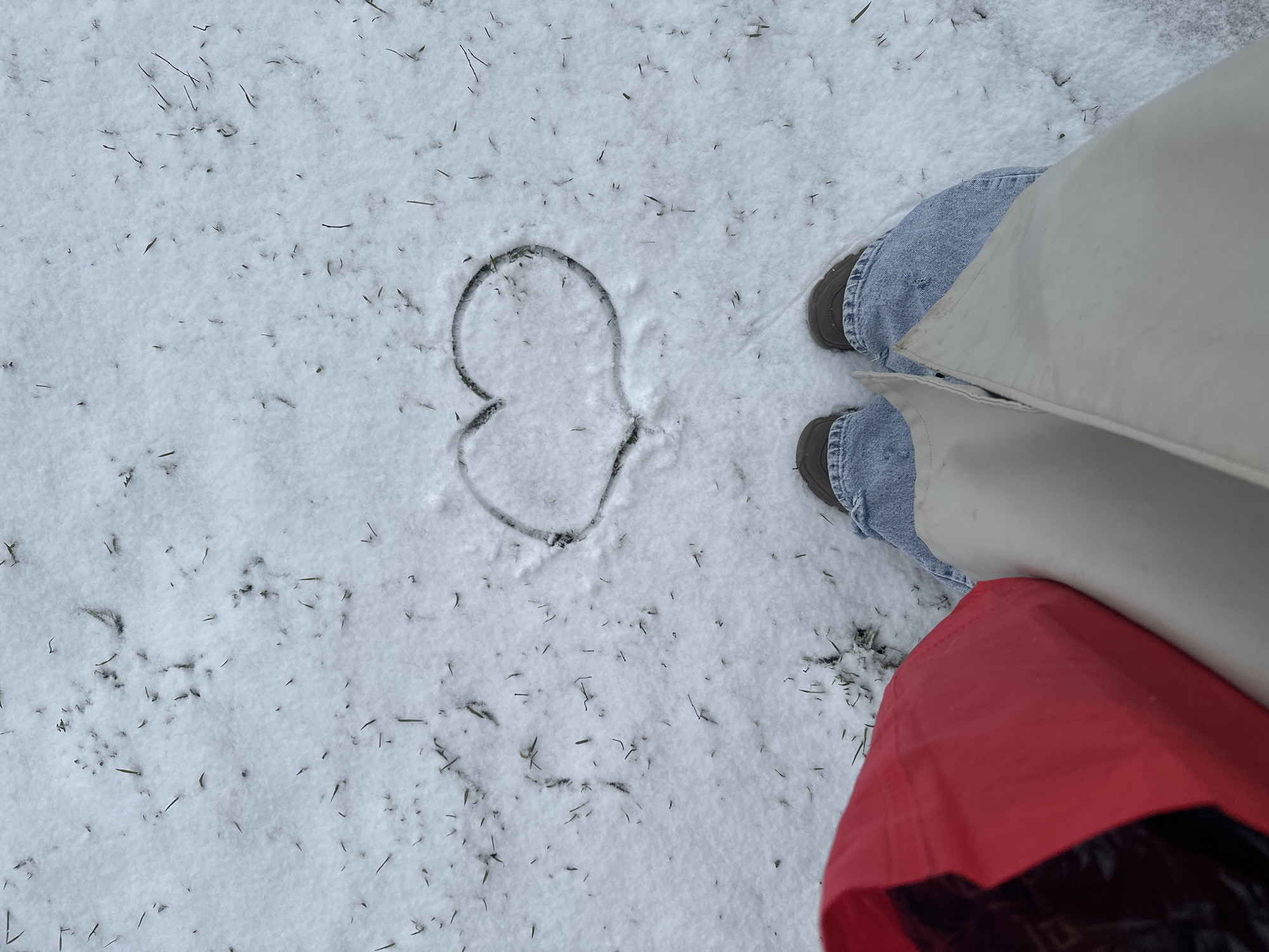 A top-down shot reveals a snow-covered ground with sparse grass visible. A crude drawing of a heart shape is imprinted in the snow. Standing near the heart are a person's feet clad in brown shoes and light-wash jeans. The person is wearing a light beige raincoat, with a portion of a red garment peeking out. There is no information available to ascertain if this image was taken in the city of Chaikovsky, Russia.