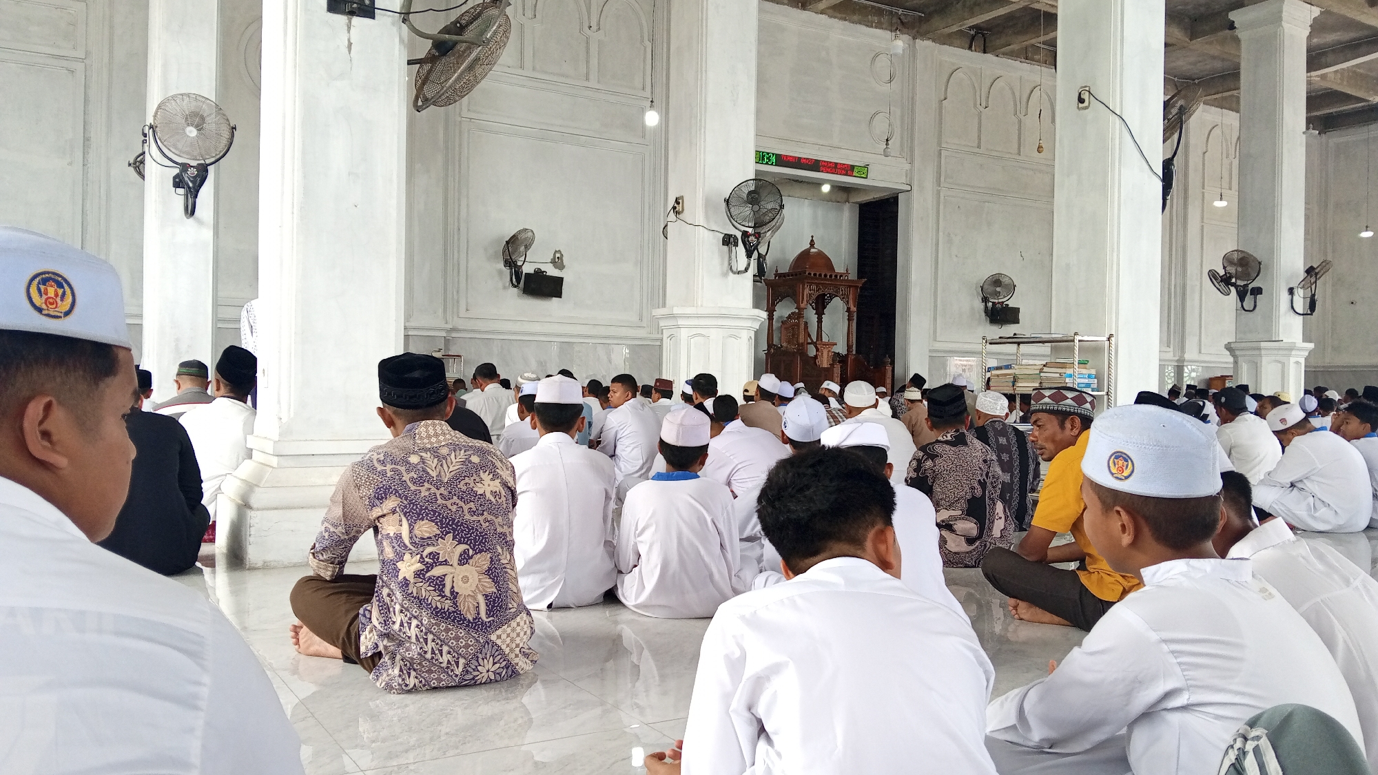 The image depicts the interior of a mosque in Bireuen, Indonesia, where a group of men and boys are seated on the floor, facing the front of the mosque. They are all dressed in white clothing, and many are wearing skullcaps or peci hats. The mosque has tall white pillars and a tiled floor that reflects the light. Fans are mounted on the walls and pillars to cool the large space. In the background, a wooden pulpit is visible, and a digital display shows time and prayer information. The men appear to be engaged in prayer or listening to a sermon. The overall atmosphere is one of reverence and community.