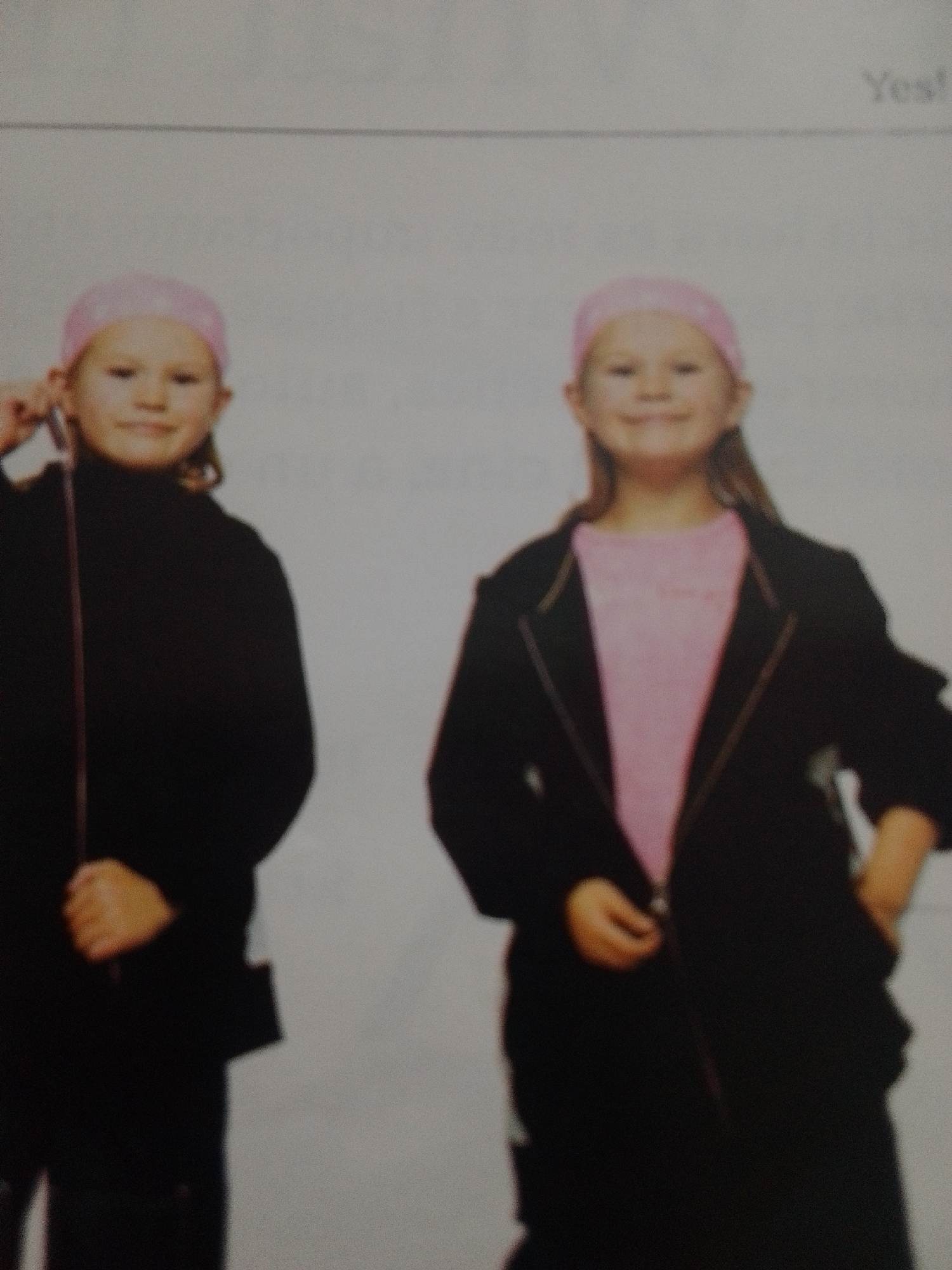Two young girls are posing for a photograph against a plain, light-colored background. Both girls are wearing pink headscarves. The girl on the left is wearing a black, long-sleeved top and is holding a thin, dark object vertically in front of her. The girl on the right is wearing a pink t-shirt and a black blazer. The image is slightly blurry and appears to be a photograph of a printed photograph, possibly from a school or personal album. There is some faint, blurry text at the top right of the image that appears to say "Yes!". There are no discernible cues about the time of day, weather, or specific location within La Tagua, Venezuela. The overall mood is neutral, as they are posing for a picture.