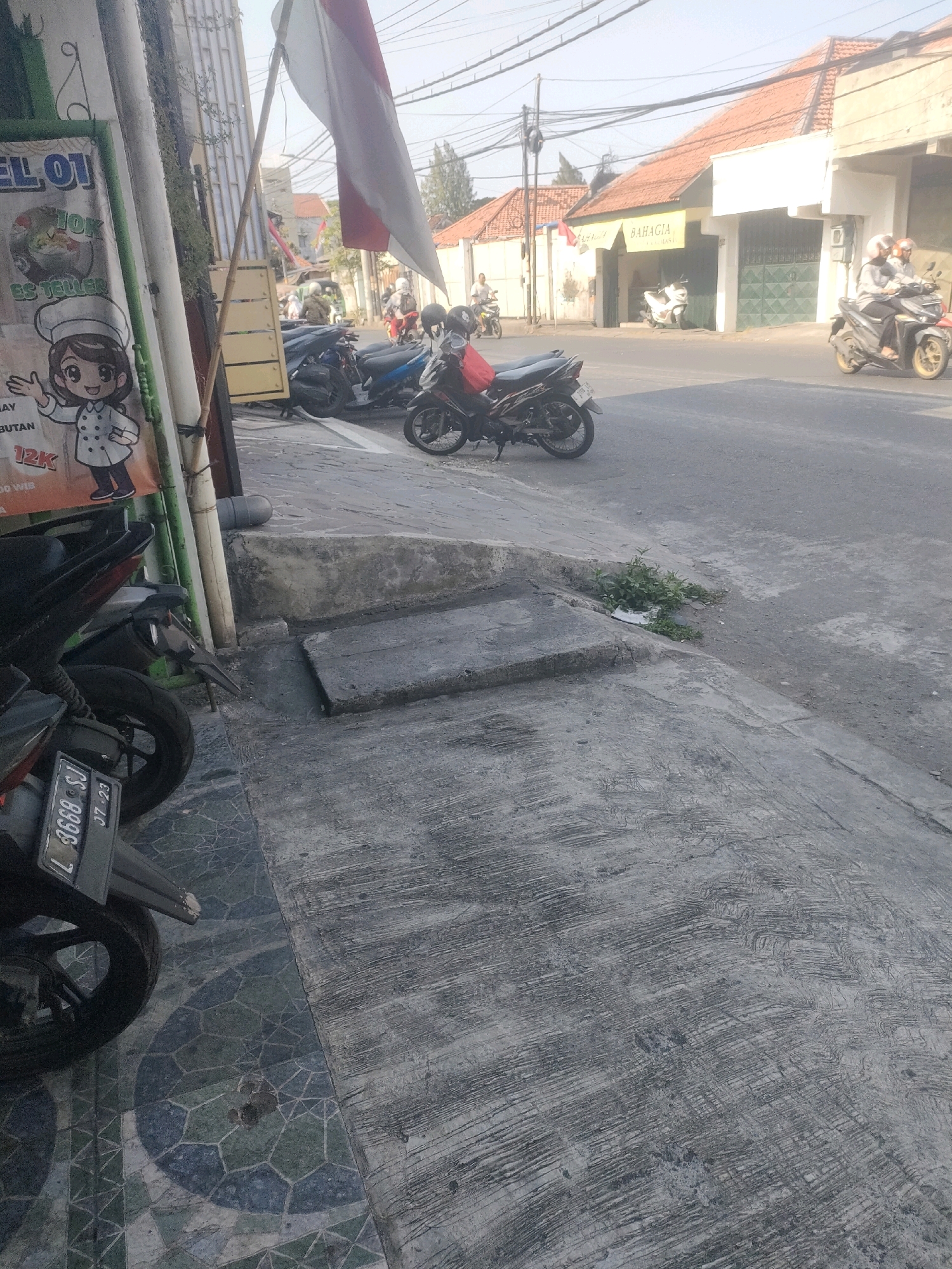 The image captures a street scene in Surabaya, Indonesia, on a clear day. In the foreground, a black motorcycle is parked on a patterned tiled pavement, with its license plate reading "L 3668 SJ 07-23". To the left, a vertical pole supports a white and red Indonesian flag. Behind the motorcycle, another black motorcycle is parked. Further back, a row of motorcycles is parked along the side of a road. On the right side of the street, two people wearing helmets are riding a yellow motorcycle, heading away from the camera. Across the street, buildings with orange tiled roofs are visible. One building has a sign that reads "BAHAGIA". The overall environment appears to be a commercial or residential street with moderate pedestrian and vehicle traffic.