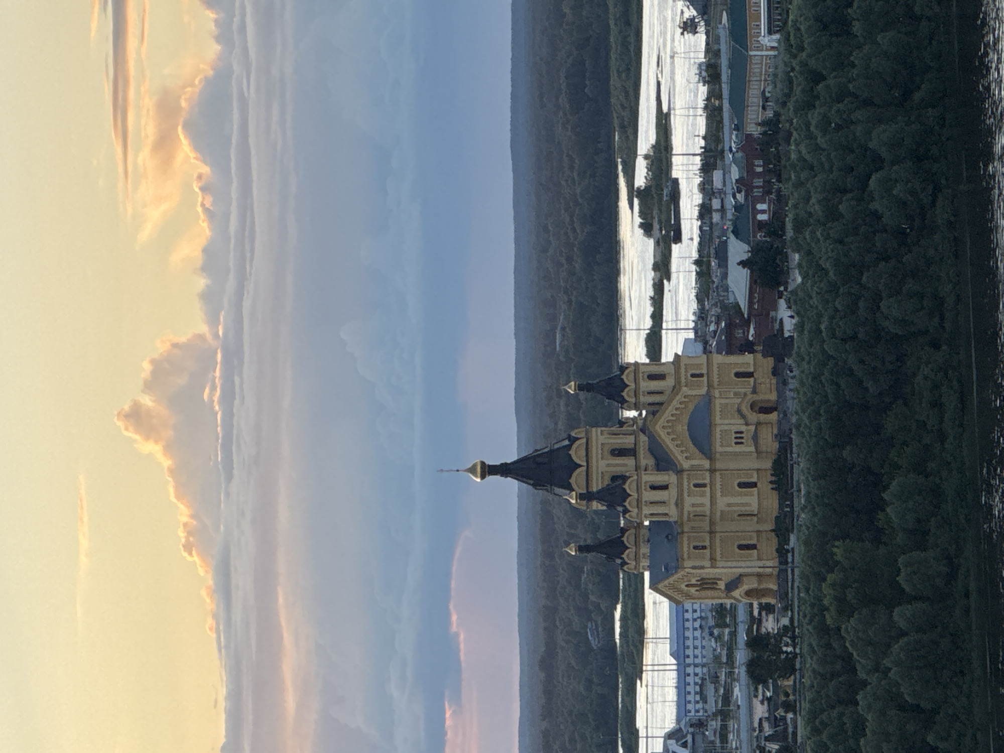 The image shows an aerial view of the Nizhny Novgorod Kremlin at sunset. The Kremlin is a historic fortress located in Nizhny Novgorod, Russia. The image captures the iconic Alexander Nevsky Cathedral, with its golden domes, as the main subject. The cathedral is surrounded by lush green trees and a body of water, likely the Volga River, reflecting the warm hues of the setting sun. The sky above is filled with dramatic clouds, some illuminated by the sunset, creating a picturesque and serene atmosphere. The architecture of the cathedral is detailed, showcasing its traditional Russian Orthodox style. The surrounding landscape suggests a vibrant city with buildings and infrastructure visible in the distance, contrasting with the natural beauty of the river and forest. The overall scene evokes a sense of historical grandeur and natural splendor.