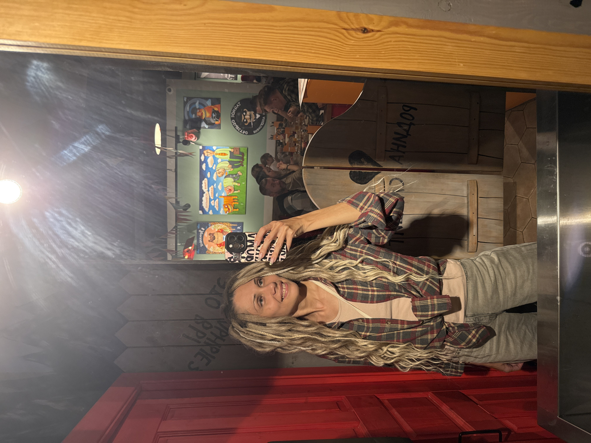 This media file is a mirror selfie taken by a woman in an indoor setting, likely a restaurant or bar, in Заволжье, Russia. The image is oriented vertically (rotated 90 degrees clockwise).
The main subject is a smiling woman with long, wavy blonde hair featuring lighter braids. She is wearing a plaid long-sleeve shirt (red, blue, and tan checks) over a light pink/beige top and light grey pants. She holds a smartphone with a black and white patterned case in her right hand to capture the reflection.
The background, visible through the mirror, shows other patrons seated at tables, appearing to dine or socialize. The walls are decorated with framed art, including a blue poster with cartoonish figures and a dark circular poster featuring a bearded character. A bright, circular light source on the left side of the mirror creates a prominent flare. The decor includes light brown wooden framing around the mirror, and wooden wall panels, some painted to resemble crates. A red door or panel is visible on the bottom left. A metallic vertical structure is on the right side of the frame.
The visible text on the circular black poster reads "ВОИН АНТИ-КОМФОРМ" (Warrior Anti-Conform). On a wooden panel in the background, "АННД09" is visible. The indoor lighting and activity suggest the photo was taken in the evening or at night. The woman's smile indicates a positive mood.