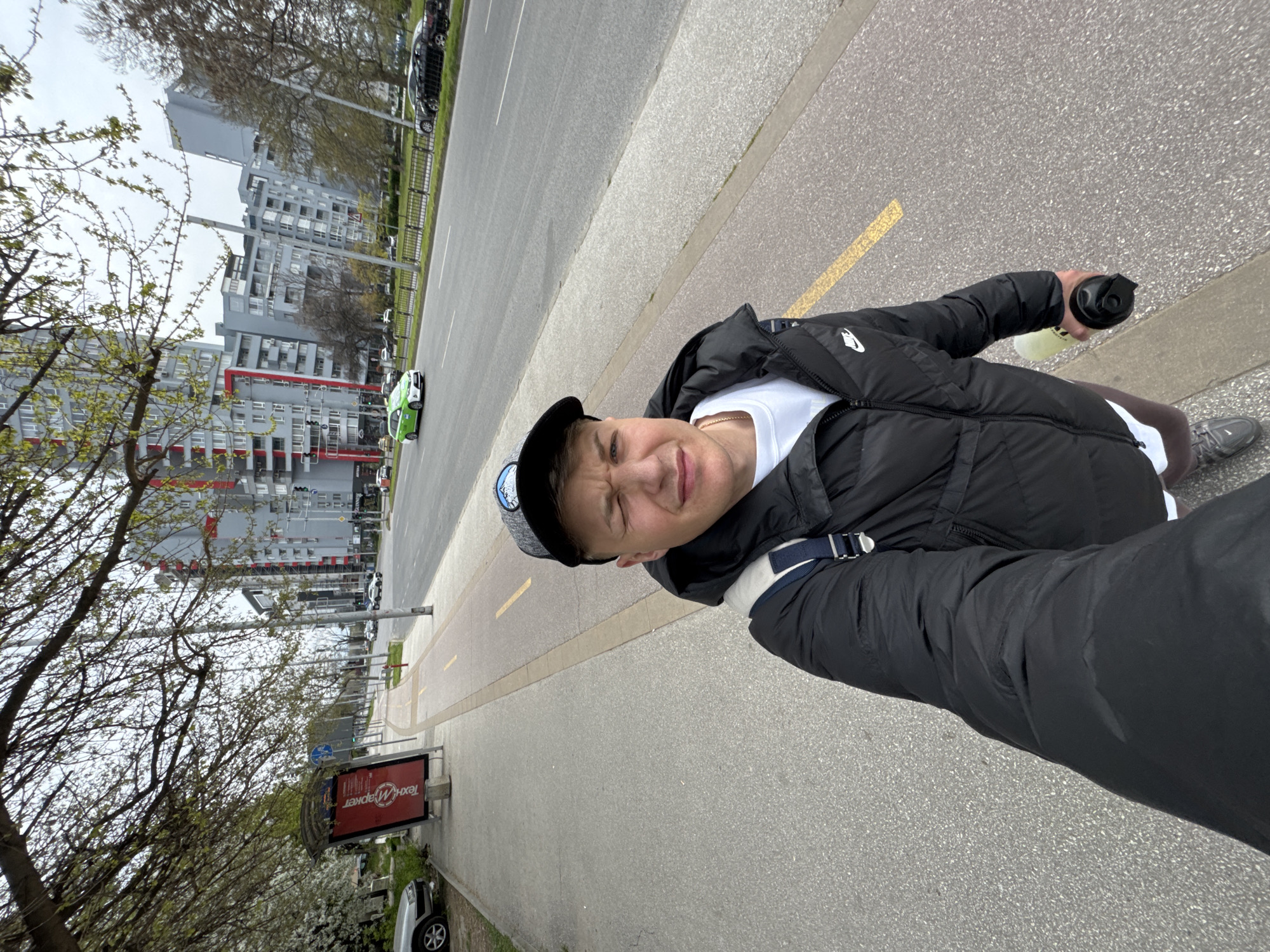 A man is walking down a paved path, likely in Plovdiv, Bulgaria. He's wearing a black jacket, a white t-shirt, and a black baseball cap. He's holding a water bottle in his right hand. He appears to be taking a selfie.
The path has a yellow painted stripe indicating a pedestrian or bike lane. To the left of the path is a sidewalk, and beyond that, a road with traffic, including a green electric car. Further away are several gray apartment buildings with red accents. There is also a bus stop sign to the left side of the image, with a “Tex Маркер” sign visible. Trees with bare branches and some with spring blossoms are visible along the sidewalk. The weather appears overcast.