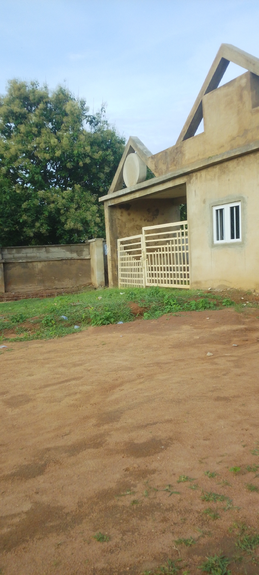 This media file depicts an outdoor scene in Jalingo, Nigeria, likely during the daytime, given the clear sky and diffused light. The primary subjects are an unfinished building with a distinctive architectural style featuring triangular and arched elements, and a light-colored metal gate. The building's facade is a neutral, sandy color, and it has a single window with white panes.
To the left of the building, there is a concrete wall, and behind it, a dense green tree with light-colored blossoms or seed pods is visible. The ground in front of the building and gate is a mix of dirt and patchy green grass. There are some plastic wrappers scattered on the grass. The overall impression is one of ongoing construction or development, with the building appearing to be in a state of partial completion.
The scene's setting cues, such as the type of vegetation, the building materials, and the overall atmosphere, suggest a location in Nigeria. The sky is a pale blue, indicating it's not a harsh midday sun, possibly early morning or late afternoon. There is no visible text in the image. The scene is devoid of people, focusing solely on the architecture and its immediate surroundings.
