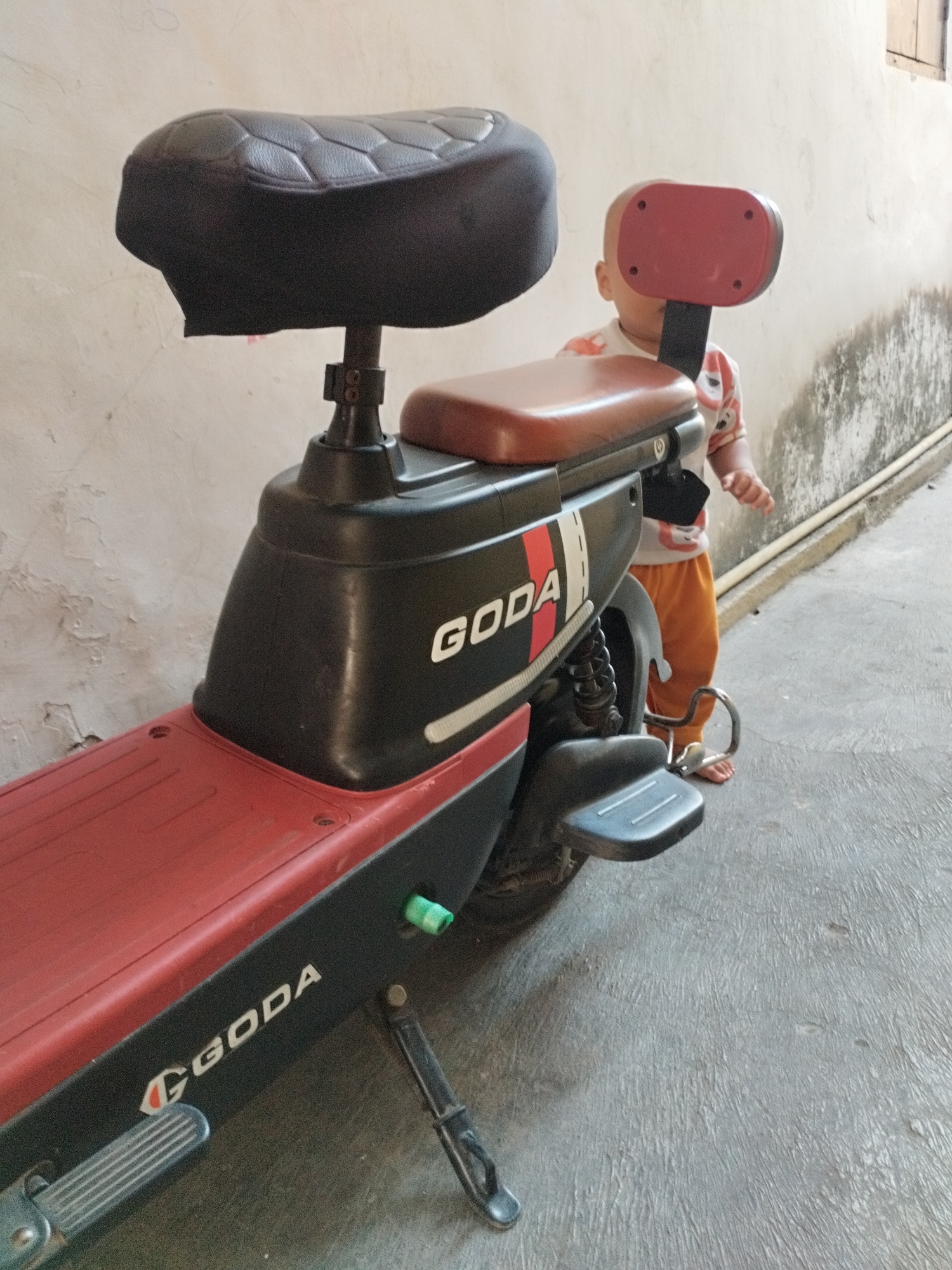 A young child stands behind a black and red electric scooter. The scooter has "GODA" printed on its side. The child is wearing an orange and white striped shirt and orange pants. The scooter is parked on a gray concrete surface next to a light-colored wall. The scene appears to be an outdoor area, possibly a residential street or courtyard, in Surabaya, Indonesia.