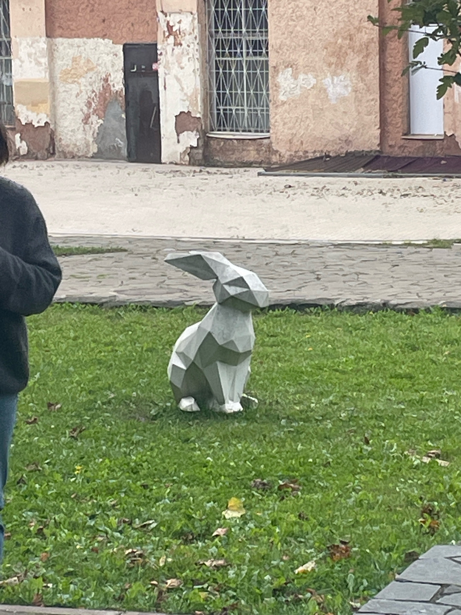 A person stands on the left, wearing a dark sweater and blue jeans. In front of the person, on a grassy area, is a faceted, geometric sculpture of a rabbit, rendered in a light gray material. The sculpture is seated and facing to the right. Behind the sculpture is a paved walkway with a light gray, rough texture. Further back, there is a building with peeling, light pink and beige walls, a dark door, and barred windows. Some green foliage is visible on the right side of the image.