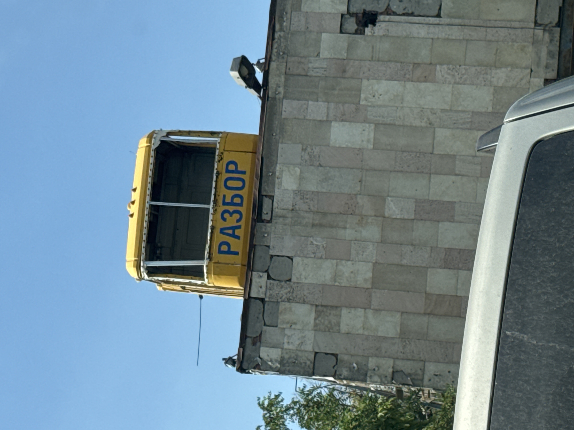 The image captures a yellow tram cabin, seemingly repurposed, mounted atop a building in Aksay, Russia. The tram cabin is positioned against a clear blue sky. On the front of the tram cabin, the word "РАЗБОР" is visible in blue Cyrillic lettering. The building beneath the tram is constructed with light-colored rectangular tiles, some of which appear to be chipped or damaged. A street lamp is affixed to the roof of the building, to the right of the tram cabin. To the left of the tram, a small, dark-colored object, possibly a bird or a small statue, is perched on the edge of the roof. In the foreground, the top portion of a silver vehicle is visible, suggesting the photograph was taken from a street level perspective. Lush green foliage from a tree is visible on the far left side of the image.