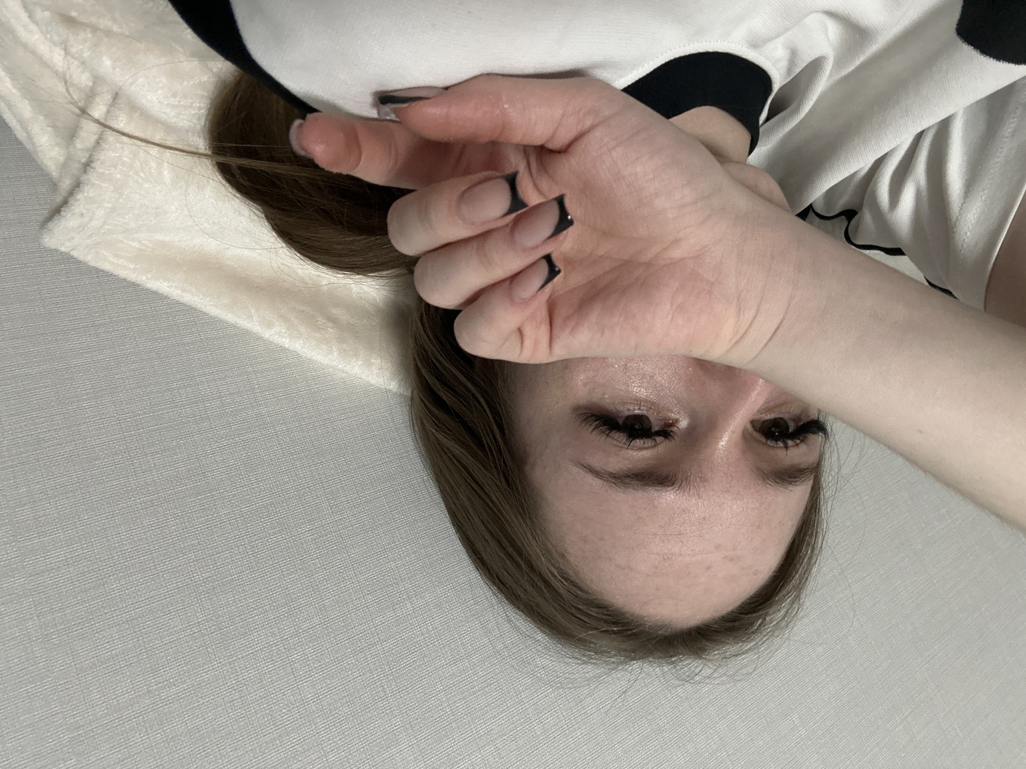 An indoor, close-up shot captures a person, likely female, lying down. The image is rotated, presenting the person's head at the bottom, appearing upside down. The subject has long, brown hair spread around their head. One hand is raised, partially obscuring the forehead and upper face. Visible features include wide-open eyes with long, dark eyelashes, and a forehead with some visible freckles and a slight sheen. The hand has manicured nails, featuring a nude base with distinct black tips in a modern French manicure style. The person is wearing a white garment with black accents, possibly a sweatshirt or long-sleeved top, parts of which are visible around the head and arm. The background consists primarily of a light gray, textured surface, possibly a wall or floor, and a soft, light-colored fabric (like a blanket or towel) is visible near the hair. No text is visible in the image.