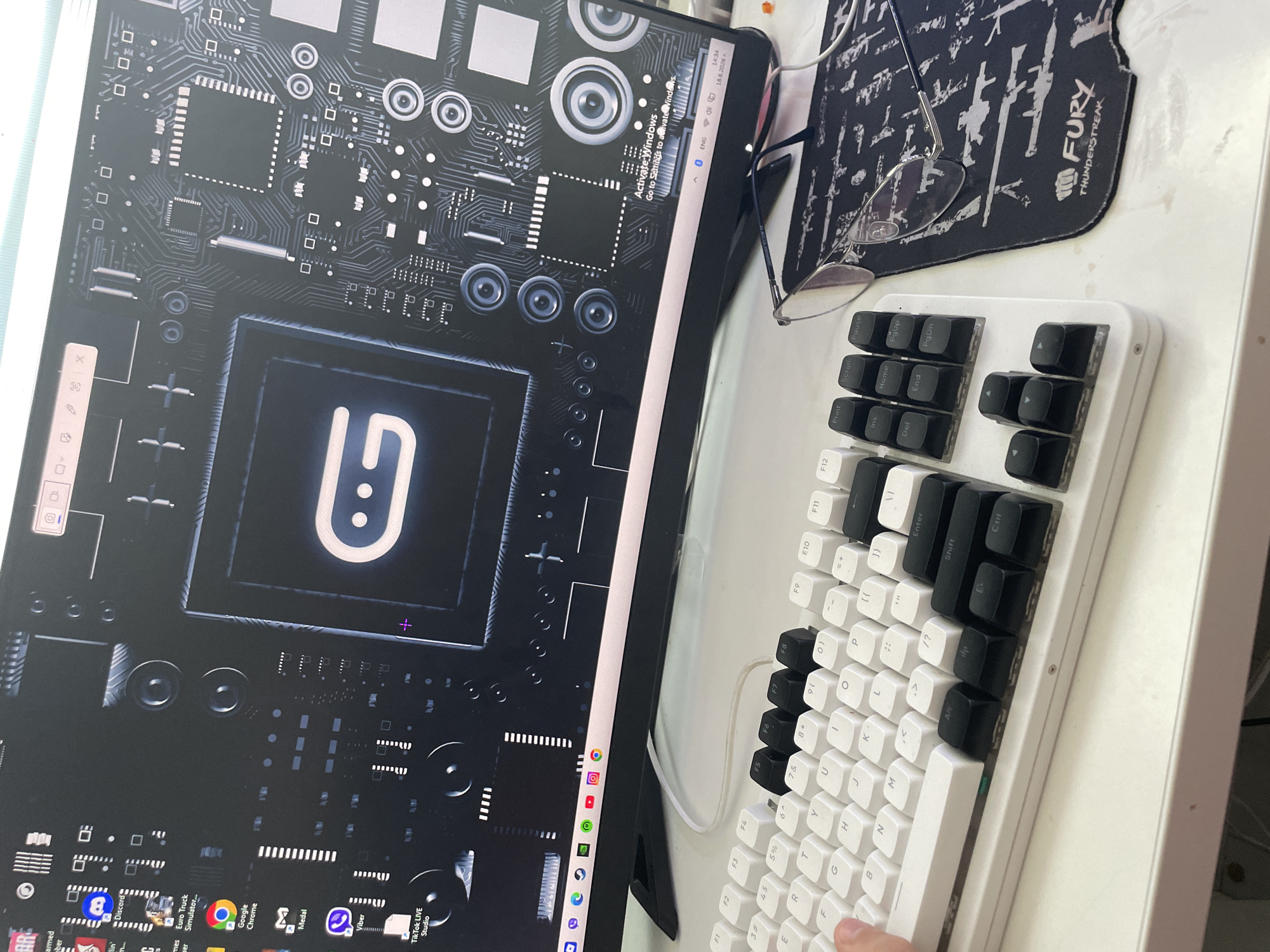 This media file is a close-up, overhead shot of a desk setup. The primary objects are a computer monitor displaying a futuristic circuit board design with a prominent, stylized "9" logo, and a white mechanical keyboard. The keyboard is partially visible, with some keys clearly labeled with standard QWERTY layout, function keys (F1-F12), and navigation keys. The monitor also shows a row of application icons at the bottom, including Discord, Google Chrome, Medal, and TikTok LIVE Studio.
In the background, a black mousepad with a stylized pattern and the text "FURY THUNDERSTREAK" is visible. A pair of eyeglasses rests on the mousepad. The overall setting appears to be a workspace, likely indoors given the artificial lighting suggested by the monitor's brightness. There's no indication of time of day or weather.
A partial view of a hand is visible on the lower right, touching the keyboard, suggesting someone is interacting with the computer. The monitor displays a "Activate Windows" prompt, indicating the operating system may not be fully licensed. The visible text includes keyboard labels, application names, and the mousepad branding. The location context provided is Petrich, Bulgaria, but there are no specific visual cues in the image to confirm this.