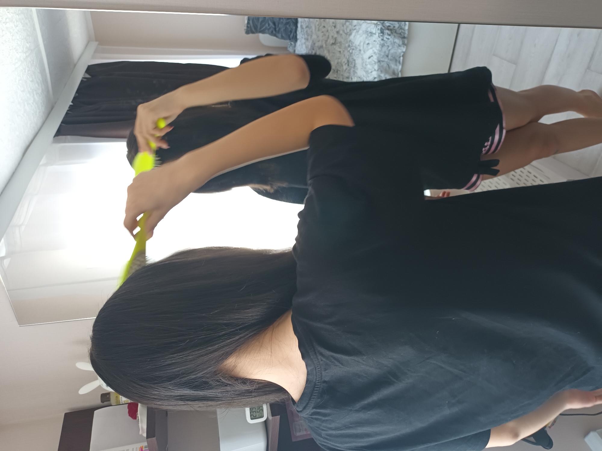 A woman with long, dark hair, wearing a black t-shirt, is brushing her hair in front of a mirror. The reflection in the mirror shows the woman's legs and a black short and pink stripes. The image includes a glimpse of a room with a patterned bedspread and light-colored flooring. On the shelf a clock is visible reading 23:54. Location: likely Ольховка, Russia.