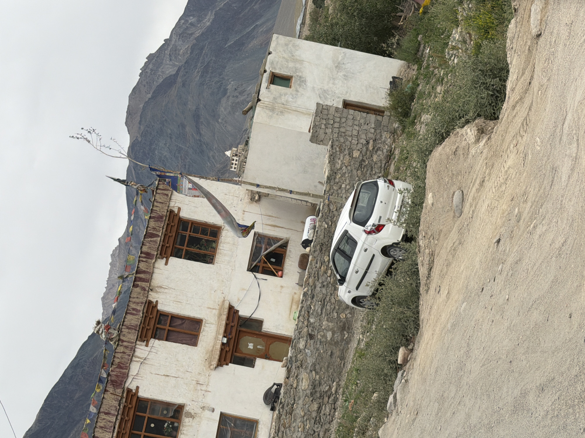 The image depicts a scene in the city of Padum, India, characterized by its mountainous terrain and traditional architecture. In the foreground, a white hatchback car is parked on a dirt road amidst sparse greenery. The car is angled slightly uphill, with its rear facing the viewer. Behind the car, a stone wall separates the road from a building.
The building itself is constructed in a traditional style, with whitewashed walls and wooden-framed windows. Prayer flags are strung along the roofline, adding a splash of color against the overcast sky. The building appears to be multi-story, with several windows visible. Further behind the primary building, another structure with a white facade and a single green-windowed section is visible, topped with a wooden beam. In the distance, a majestic mountain range dominates the skyline, its rugged slopes conveying a sense of remoteness and grandeur. The sky is heavily clouded, suggesting a potentially cool or damp atmosphere. The overall impression is one of a serene, somewhat austere, yet culturally rich environment set against a dramatic natural backdrop.