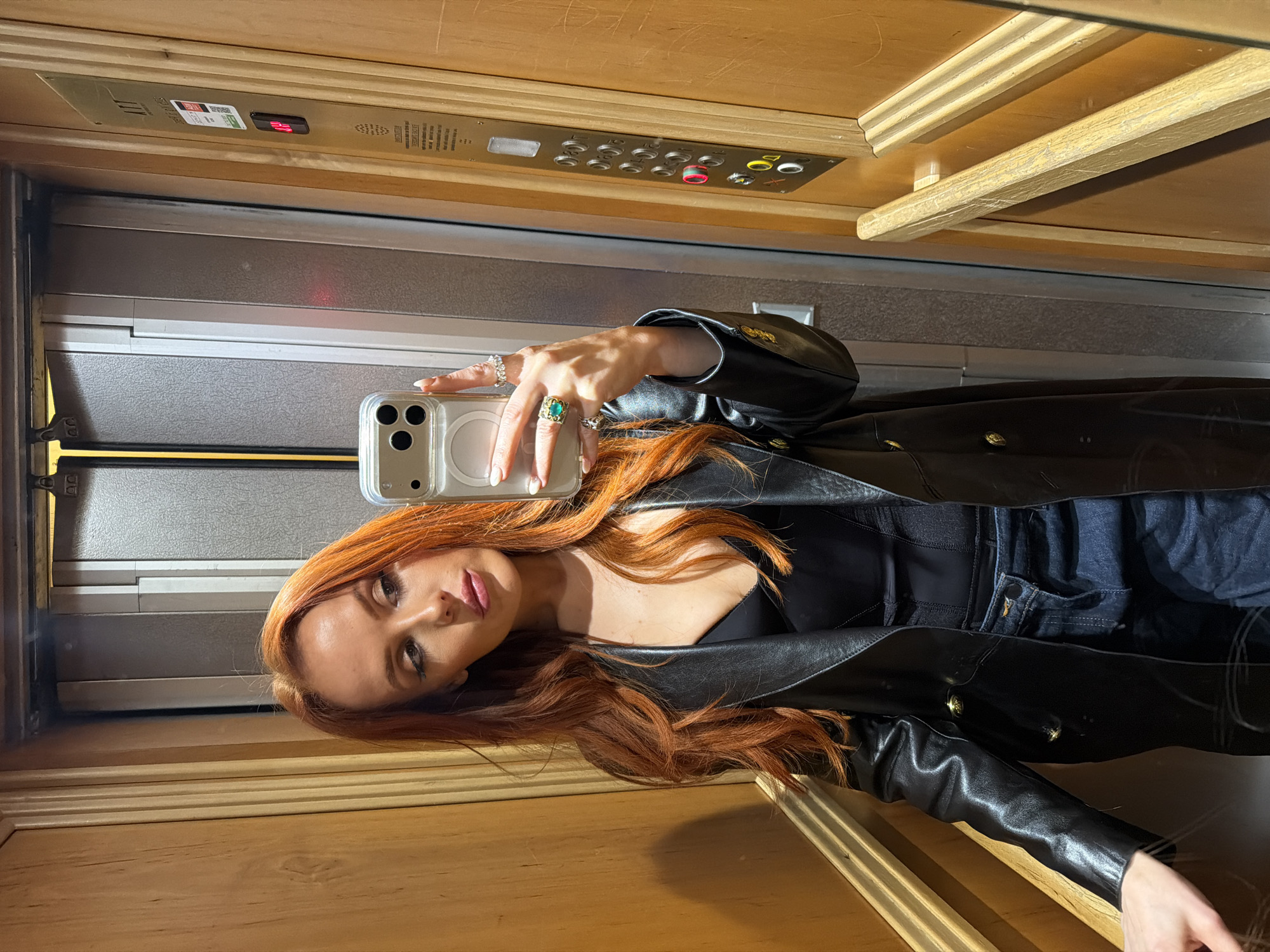 A woman with red hair is taking a selfie in an elevator with wooden walls and a brushed metal door. She is wearing a black leather jacket, a black top, and dark denim jeans. She is holding an iPhone in her left hand, which is adorned with multiple rings, including one with a large green stone. The elevator control panel is visible in the background. The woman's location is not discernible from the image.