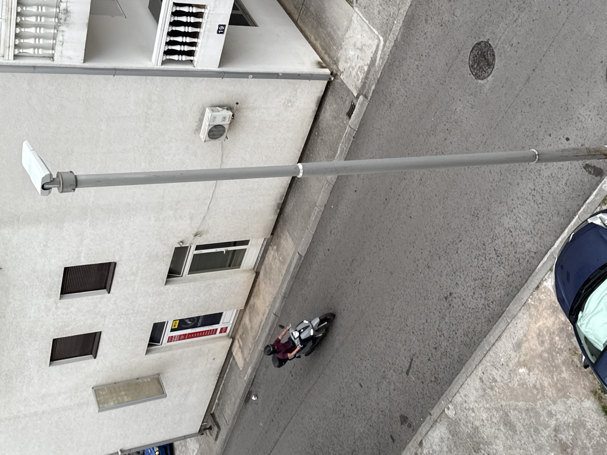 The image, taken from an elevated perspective, depicts a street scene in Budva, Montenegro. A light gray multi-story building stands to the left, featuring white facade, multiple dark windows, and a light gray streetlamp extending over the road. The number "19" is visible on the building. An air conditioning unit is mounted on the building's facade.
A motorcyclist wearing a maroon top and a helmet is riding on the street next to the building. A blue car is parked on the right side of the road. A manhole cover is visible on the road surface. The asphalt road shows signs of wear and tear.