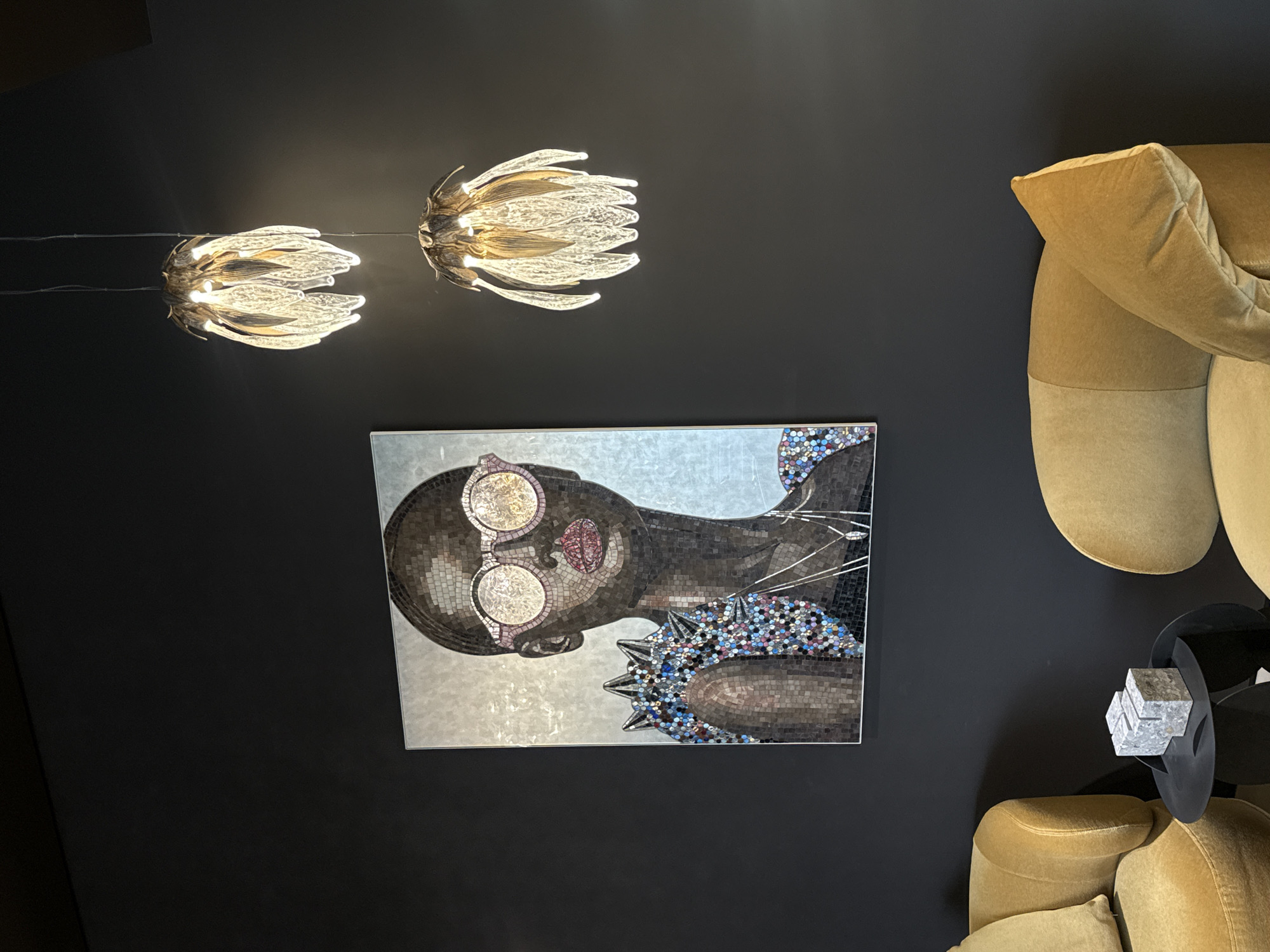 The image displays a sophisticated interior space, likely a living room or lounge area in Moscow, Russia, characterized by a dramatic dark grey or black wall. Two elegant ceiling lights hang from above, fashioned from textured glass or acrylic in a warm, flower-like or organic design, emitting a soft, inviting glow.
A prominent feature is a large rectangular mosaic artwork on the wall, depicting the upper body of a stylized person, possibly a woman, crafted entirely from mosaic tiles. The figure has dark skin tones and is shown in profile, adorned with distinctive round glasses made from shimmering, light-colored mosaic pieces. Her lips are a contrasting reddish-pink, and her shoulder and chest area are decorated with a vibrant, multi-colored mosaic pattern that includes intricate, spiky details. The background of the mosaic is a reflective silver or light grey.
To the right of the artwork, a plush, comfortable sofa or armchair, upholstered in a warm mustard yellow or light brown fabric, features soft, rounded forms. A small, elegant black circular side table is positioned between sections of the seating furniture. On this table rests a unique decorative object composed of several stacked, irregular silver or grey cubes, adding a modern, sculptural touch to the room's decor.
There are no actual people present, only the mosaic representation. The indoor lighting suggests a serene ambiance, possibly in the evening. No visible text is present in the scene. The overall impression is one of artistic taste and comfortable luxury.