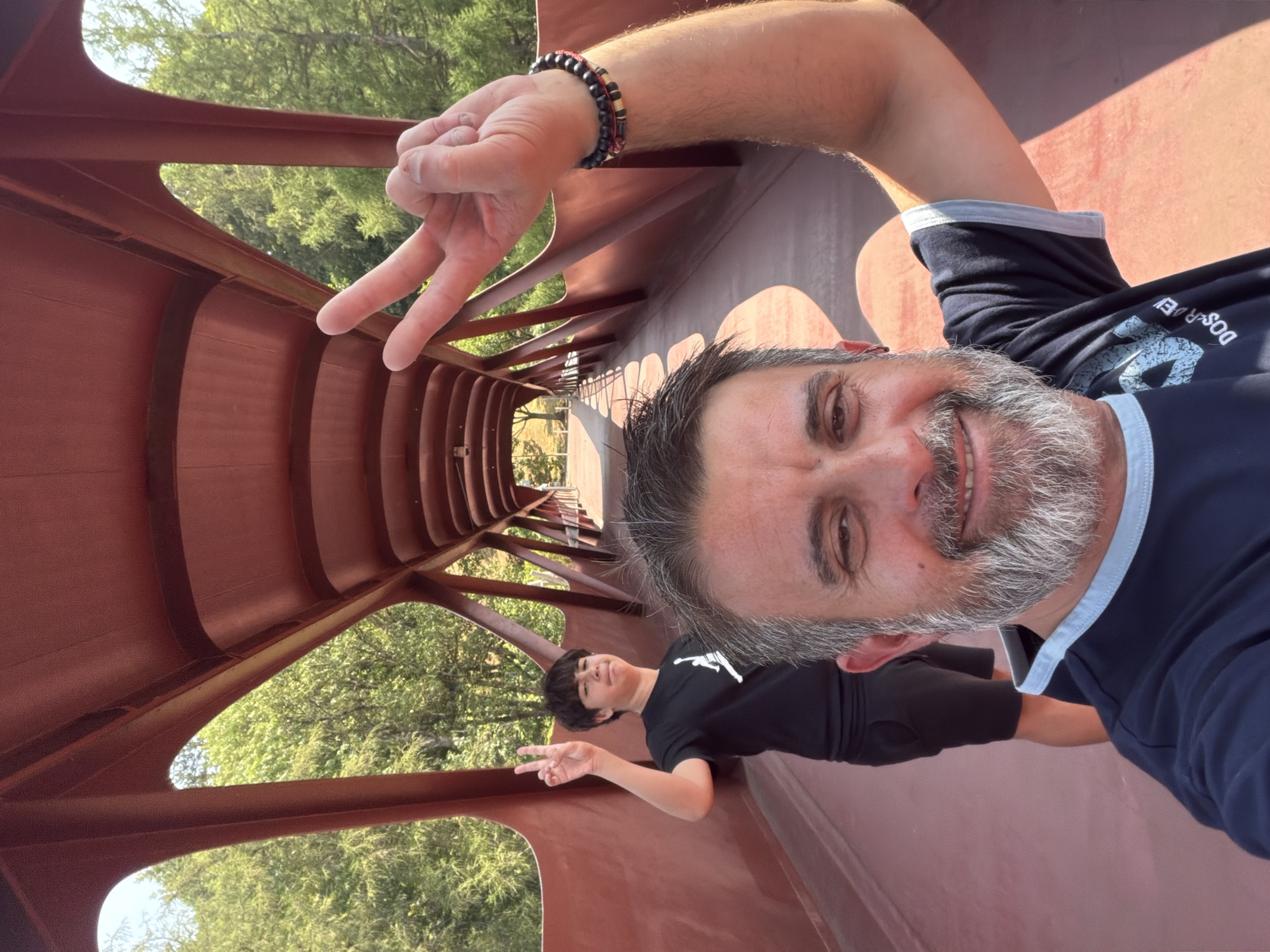 Two males are on a pedestrian bridge. The male in the foreground is taking a selfie, smiling and holding up a peace sign with his right hand. He has a grey beard and is wearing a dark blue t-shirt with a light blue collar. He is also wearing bracelets on his right wrist. The male in the background is standing behind the first male and to his left. He is also holding up a peace sign with his left hand and is wearing a black t-shirt and black shorts. The bridge is made of red metal and has a covered walkway. Trees and foliage are visible on either side of the bridge. This bridge is the Passerelle Leonardo da Vinci, located in Esch-sur-Alzette, Luxembourg.