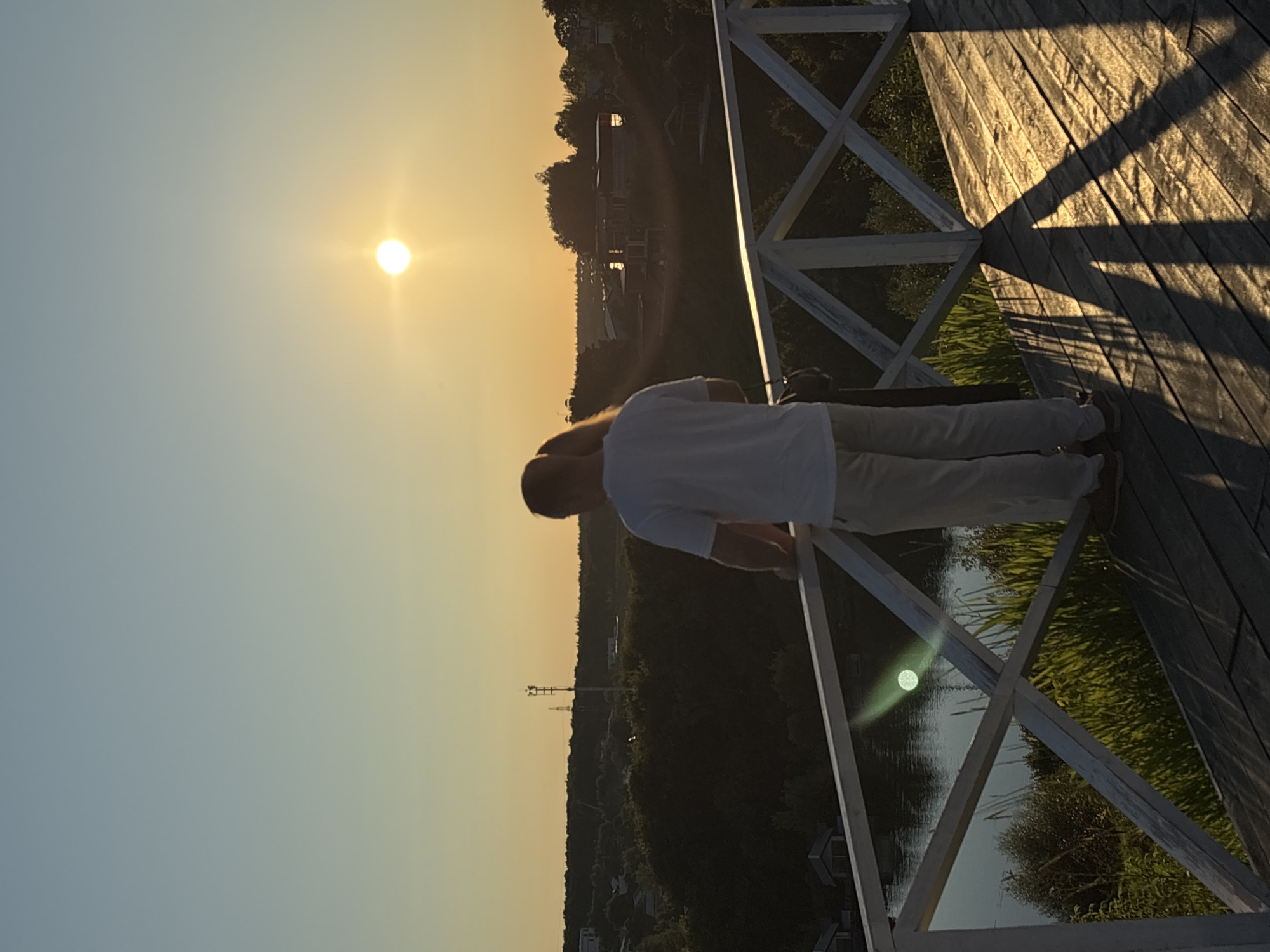 A man and a woman stand on a wooden platform overlooking a body of water and a tree-lined landscape. The sun is setting in the background, casting a warm glow and long shadows across the wooden planks. The man, wearing a white t-shirt and light-colored pants, stands with his back to the camera, looking out at the view. The woman, partially visible beside him, has blonde hair. A white wooden railing with a crisscross pattern separates the platform from the water. In the distance, several houses and a cellular tower are visible amidst the trees. The scene appears to be located in Romashkovo, Russia, given the architectural style and natural surroundings.