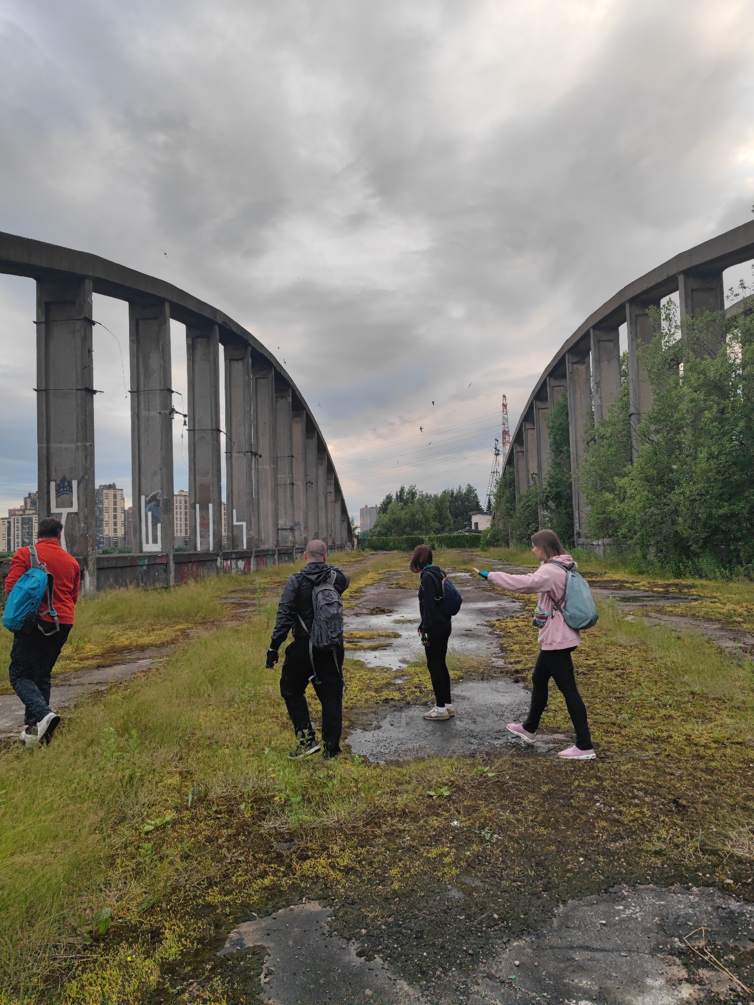 Four individuals are walking on an overgrown, abandoned bridge structure under a cloudy sky in a city setting. The bridge is characterized by large concrete pillars forming arches. In the background, tall buildings are visible, suggesting an urban environment. One person on the left wears a red jacket and a blue backpack, walking away from the camera. A person in a black jacket and backpack is in front of them, also walking away. Two people are in front of them, facing towards the right of the frame. One is wearing a dark jacket and a backpack, and the other is wearing a pink hooded jacket and a light blue backpack, with their arm extended as if pointing or interacting. The ground is covered with moss and patches of asphalt or concrete. A tall industrial crane can be seen in the distance on the right side of the image.