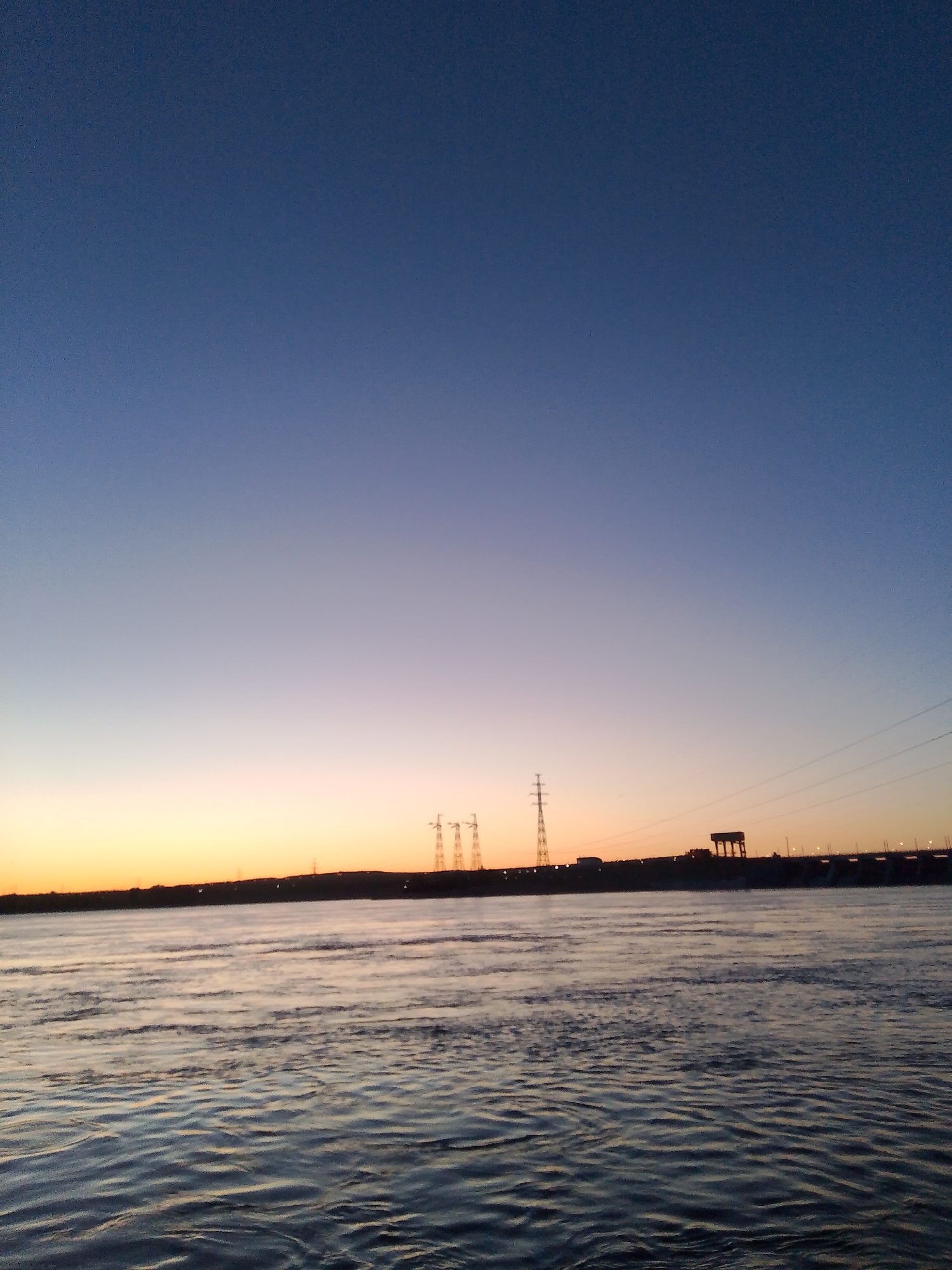 The image depicts a twilight or pre-dawn scene with a body of water in the foreground, likely a river or large lake, based on the visible waves and ripples. The water's surface is illuminated by the faint light.
In the middle ground, a dark, silhouetted landmass stretches across the horizon. Several transmission towers or power pylons are visible against the brightening sky. A structure resembling a dam or bridge is also present, with architectural features that suggest an industrial or infrastructural purpose.
The sky transitions from dark blue at the top to a lighter orange/yellow near the horizon, indicating sunrise or sunset.
Based on the location of the image, it is likely the Kama River or a large reservoir near Chaikovsky, Russia. The structure in the background could be associated with a hydroelectric power plant or a similar water management system.