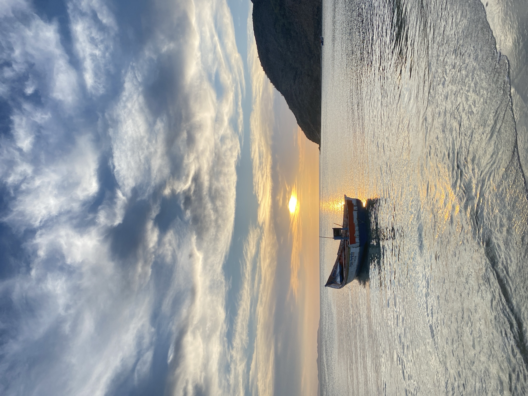 This tranquil landscape image, set in Juan Griego, Venezuela, captures a scenic moment during either sunrise or sunset. A small, traditional wooden fishing boat, painted blue and white with red accents, floats calmly on the gently rippling water. The boat is positioned slightly to the right of the frame's center.
To the right, a large, dark mountain or hill creates a prominent silhouette against the glowing horizon. The sun, low in the sky, casts a brilliant golden light that stretches across the water, creating a shimmering path of reflections. The sky above is adorned with dramatic, streaky clouds, painted in hues of blue, white, and warm gold, transitioning from cooler tones higher up to the warmer light near the horizon.
The overall atmosphere is one of serene beauty and peaceful solitude. On the side of the boat, white lettering clearly displays "Sabor del Cielo," which translates to "Taste of Heaven." Below this, "SAABEN YUAN GRIEGO" (likely Juan Griego) can be discerned, along with a registration number "P.V. 000574." The image beautifully showcases the natural charm of the Venezuelan coast at a magical hour.