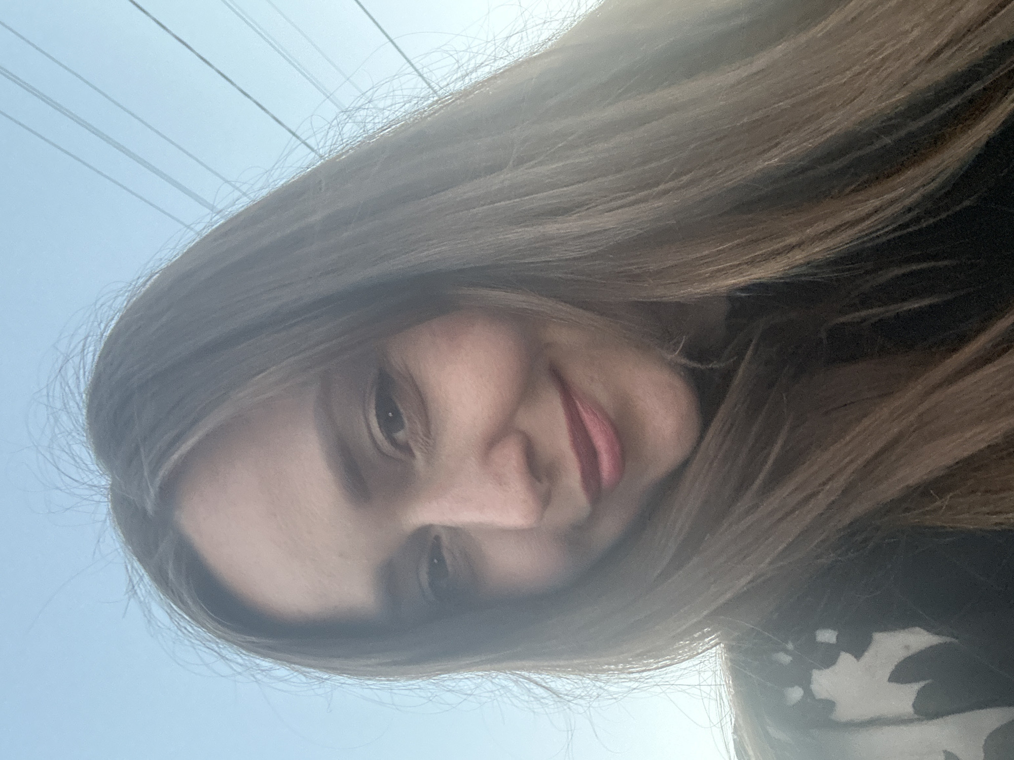 A close-up selfie of a woman with long, brown hair against a bright sky with power lines visible. She is wearing a dark shirt with a white floral pattern. Her facial features include brown eyes and pink lipstick. The lighting suggests it is daytime. The location is not specified in the image.