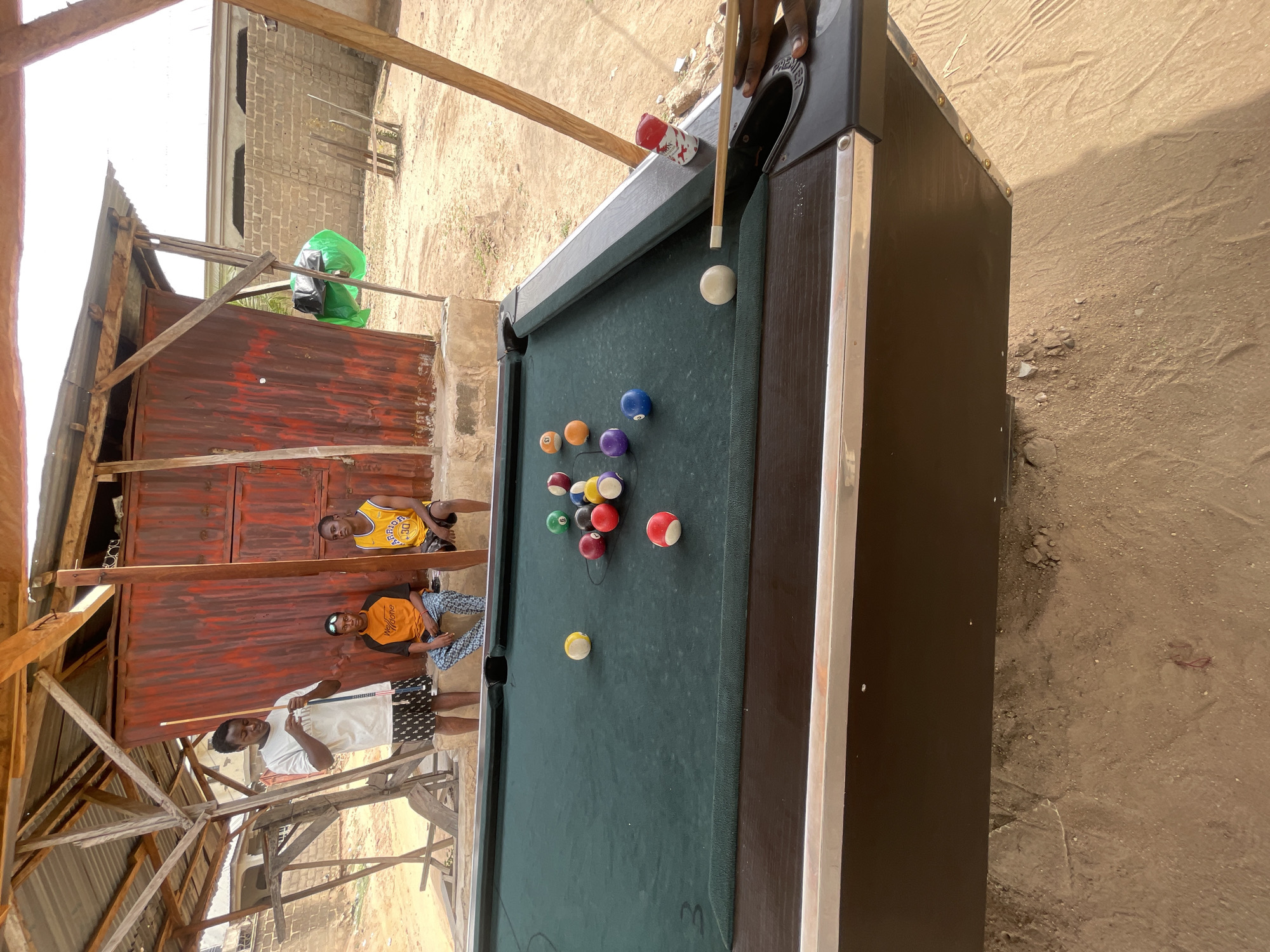 The scene is outdoors in Lokoja, Nigeria, featuring a pool table with balls arranged for a game. A person is holding a cue stick, seemingly about to make a shot. Three individuals are standing near the pool table, observing or interacting with the game. The environment includes a weathered, partially constructed shelter with wooden beams and corrugated metal walls. There's a building with exposed brick and a green object near the shelter. The ground is sandy.