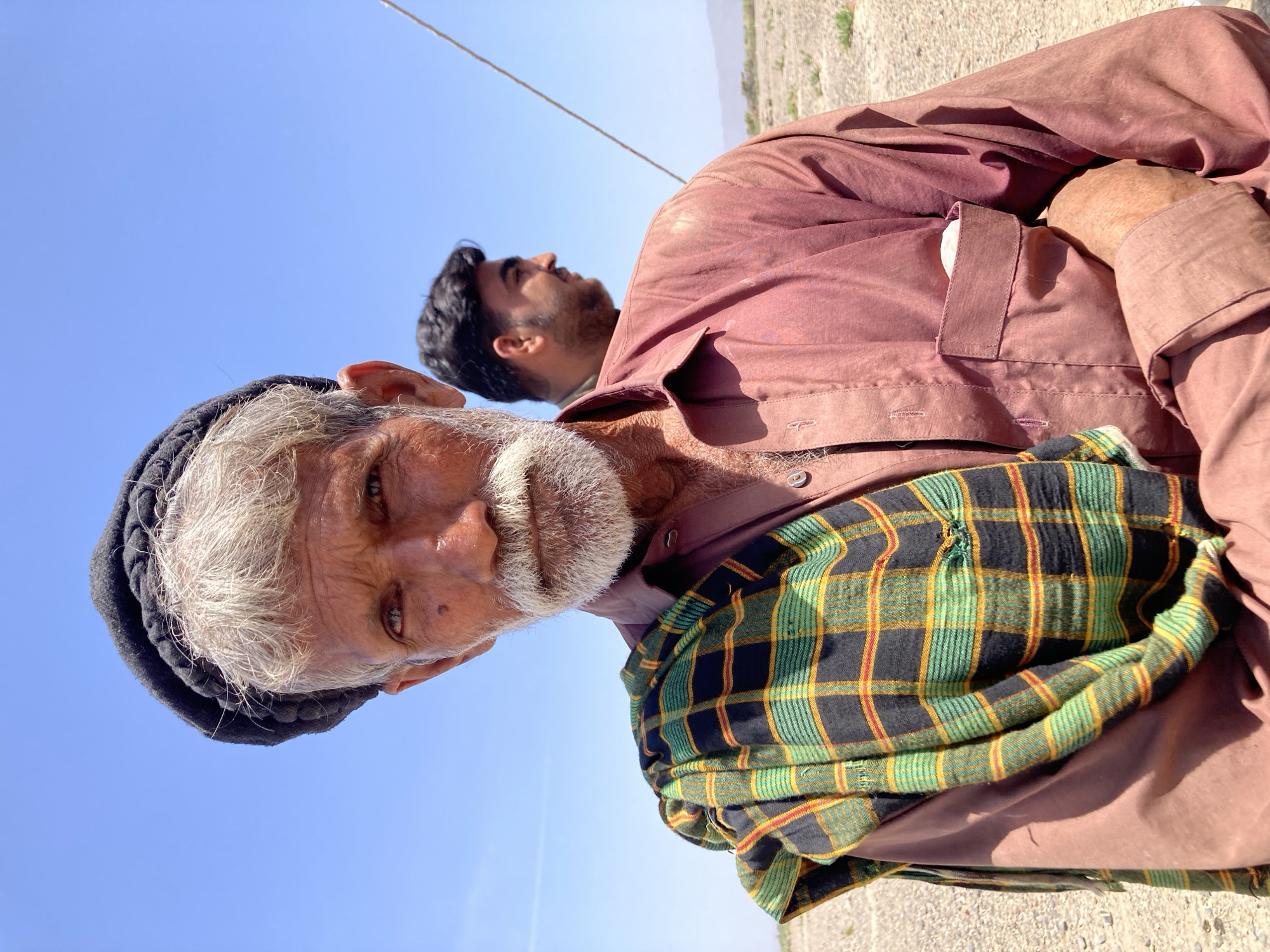 A low-angle, outdoor shot depicts an older man with a white beard and weathered skin, wearing a black head covering and a plaid shawl over a maroon shirt. The plaid pattern includes green, yellow, black, and red lines. A younger man with black hair is visible behind the older man. The background consists of a clear blue sky and a beige, dirt-covered ground. A thin cable stretches across the top of the image. The location appears to be Hussenāni, Pakistan.
